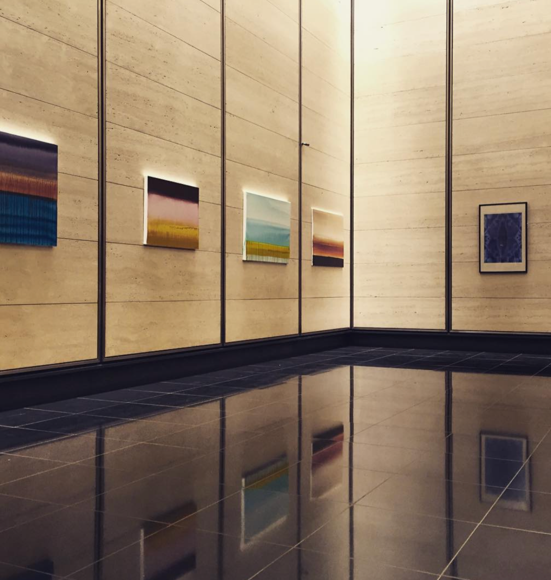 Art gallery with several colorful abstract paintings on a beige wall, reflected on the shiny black tiled floor.