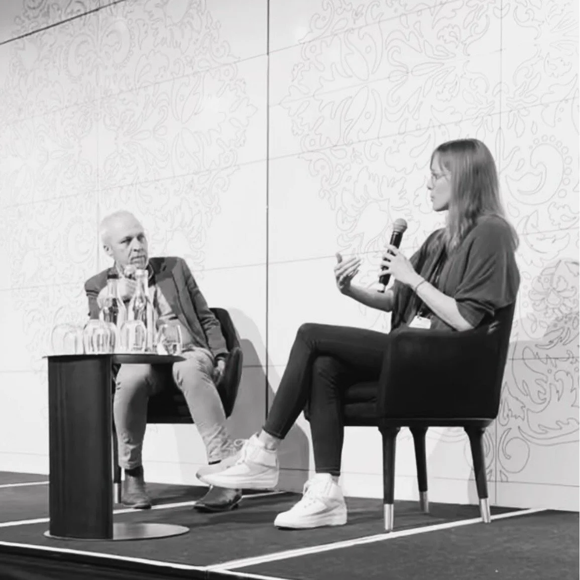 A woman and an older man seated in chairs having a conversation during a panel. The woman is holding a microphone and gesturing with her hand. The man is listening attentively. There is a small table with several water bottles in front of them, and the background features a decorative wall with floral patterns.