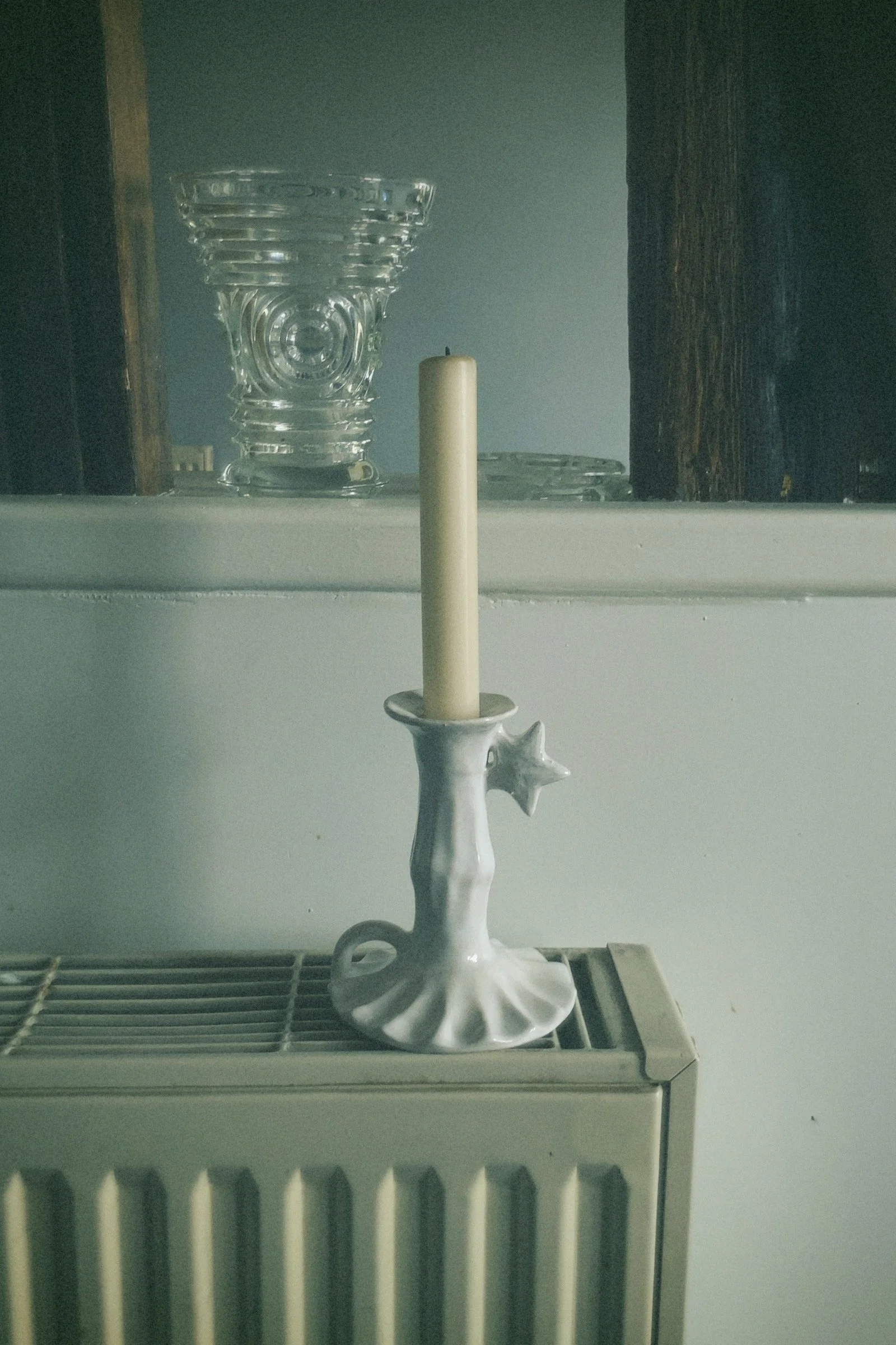 Une chandelle blanche dans un bougeoir blanc placé sur un radiateur blanc, avec un fond de porte en bois et un mur gris clair.