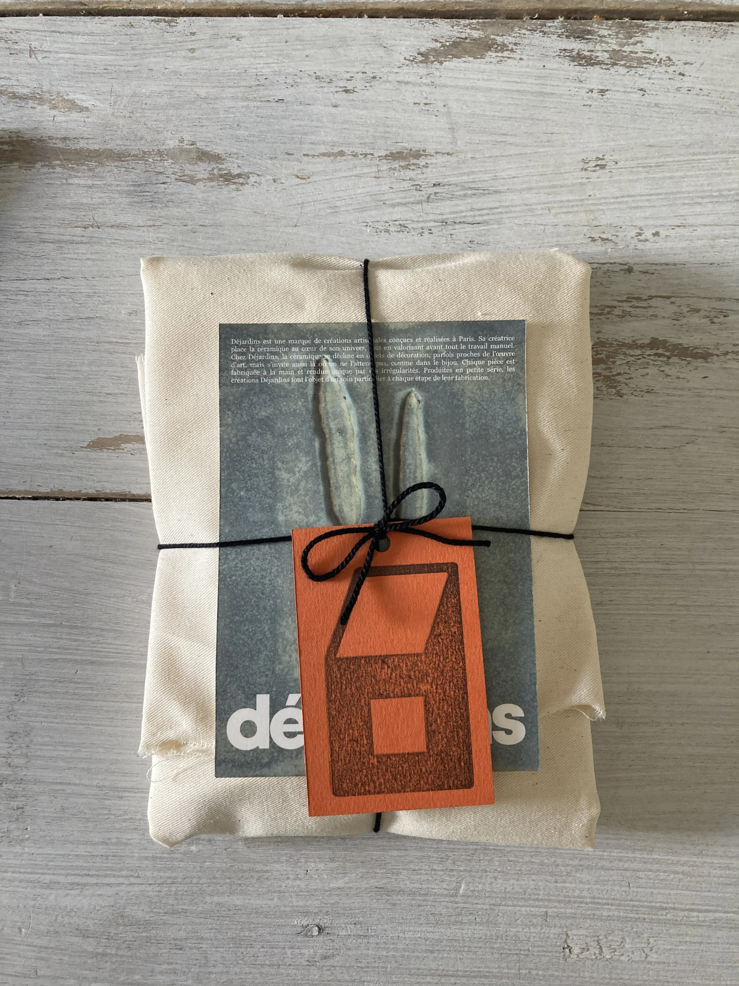 Paquet en tissu avec une carte attachée par un ruban noir, sur une surface en bois clair.