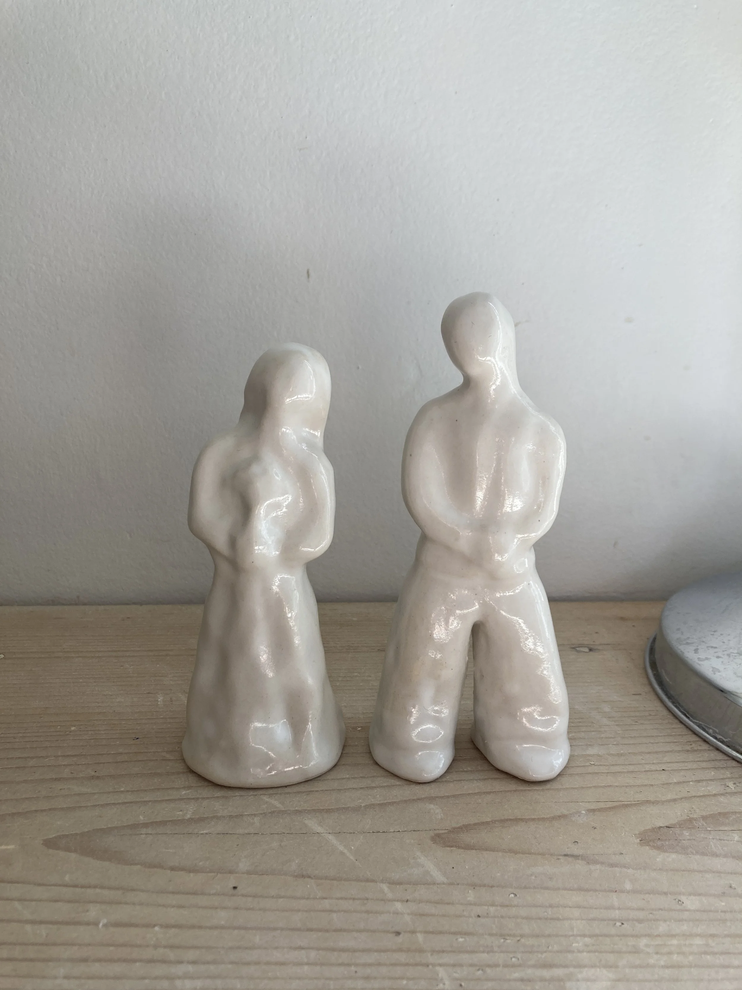 Deux sculptures en céramique blanches représentant des figures humaines sans détails faciaux, placées sur une surface en bois contre un mur blanc.