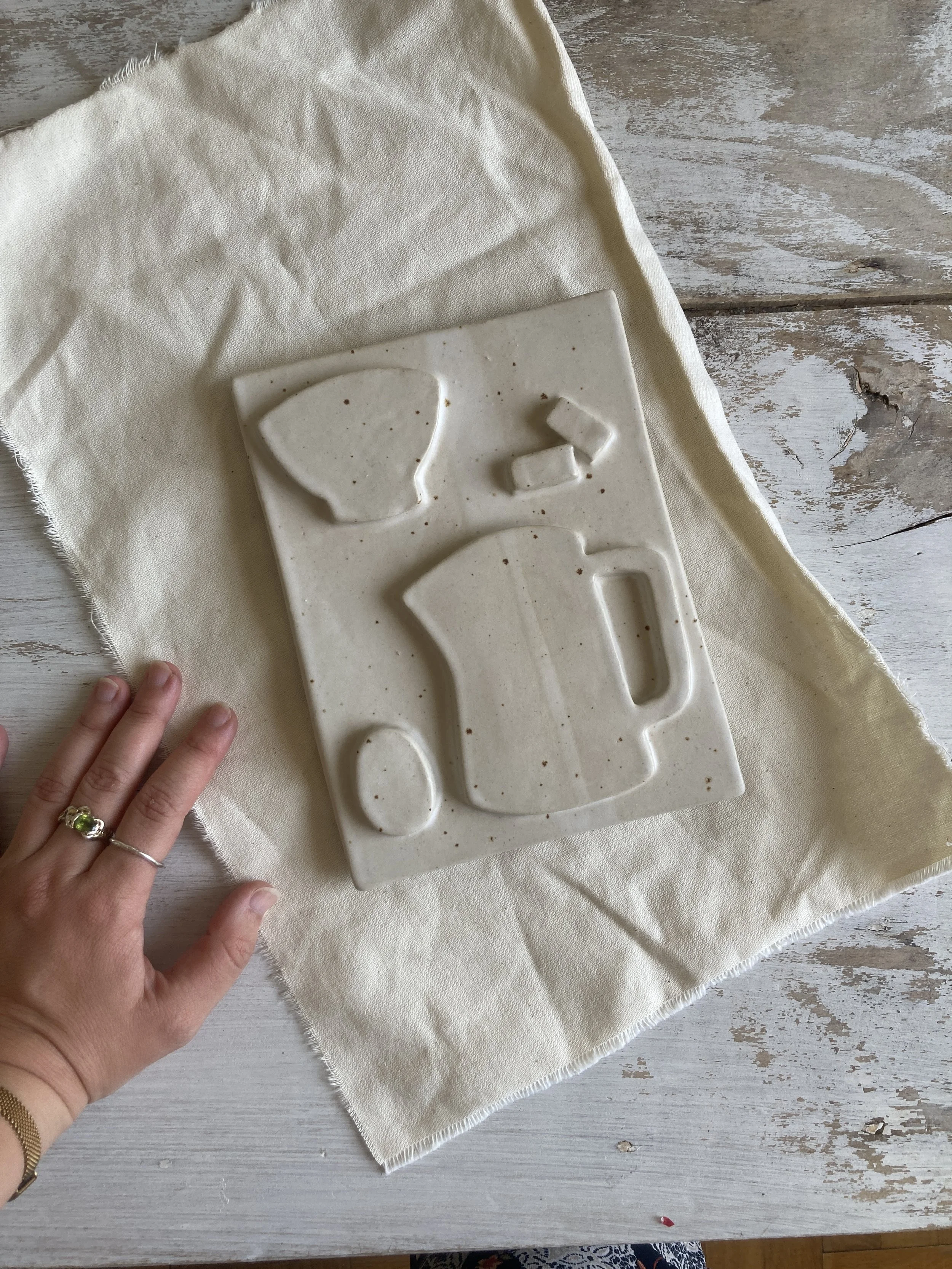 Une main tenant une plaque bas-relief en céramique blanc avec des formes géométriques comme une carafe et un bol, posée sur une nappe beige en tissu, sur une table en bois clair.