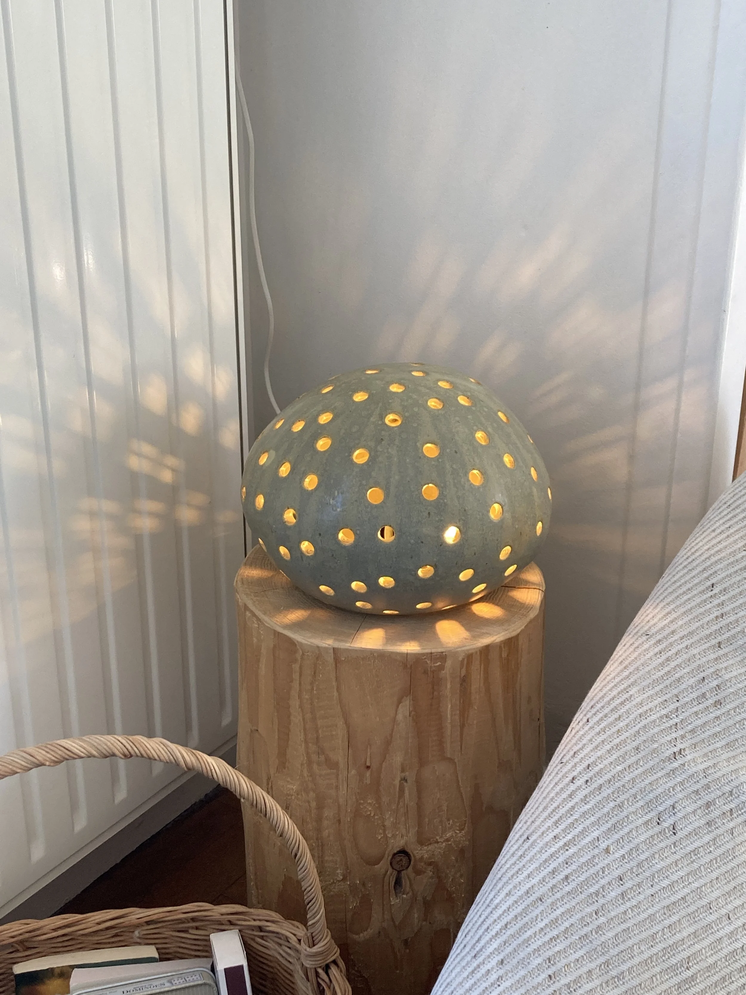 Lampe en céramique diffusant une lumière chaude avec des motifs de trous laissant passer la lumière, posée sur une table en bois, à côté d'une assise en tissu.