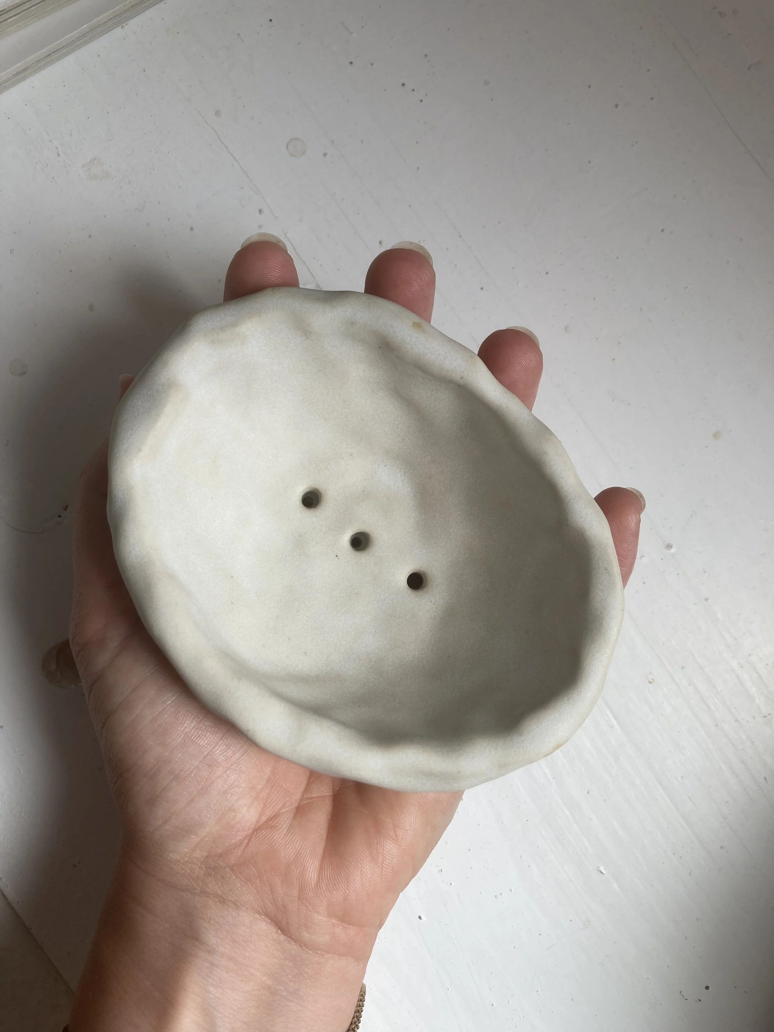 Vue du dessous d'un porte-savon en céramique blanc, tenue par une main, avec un fond blanc.