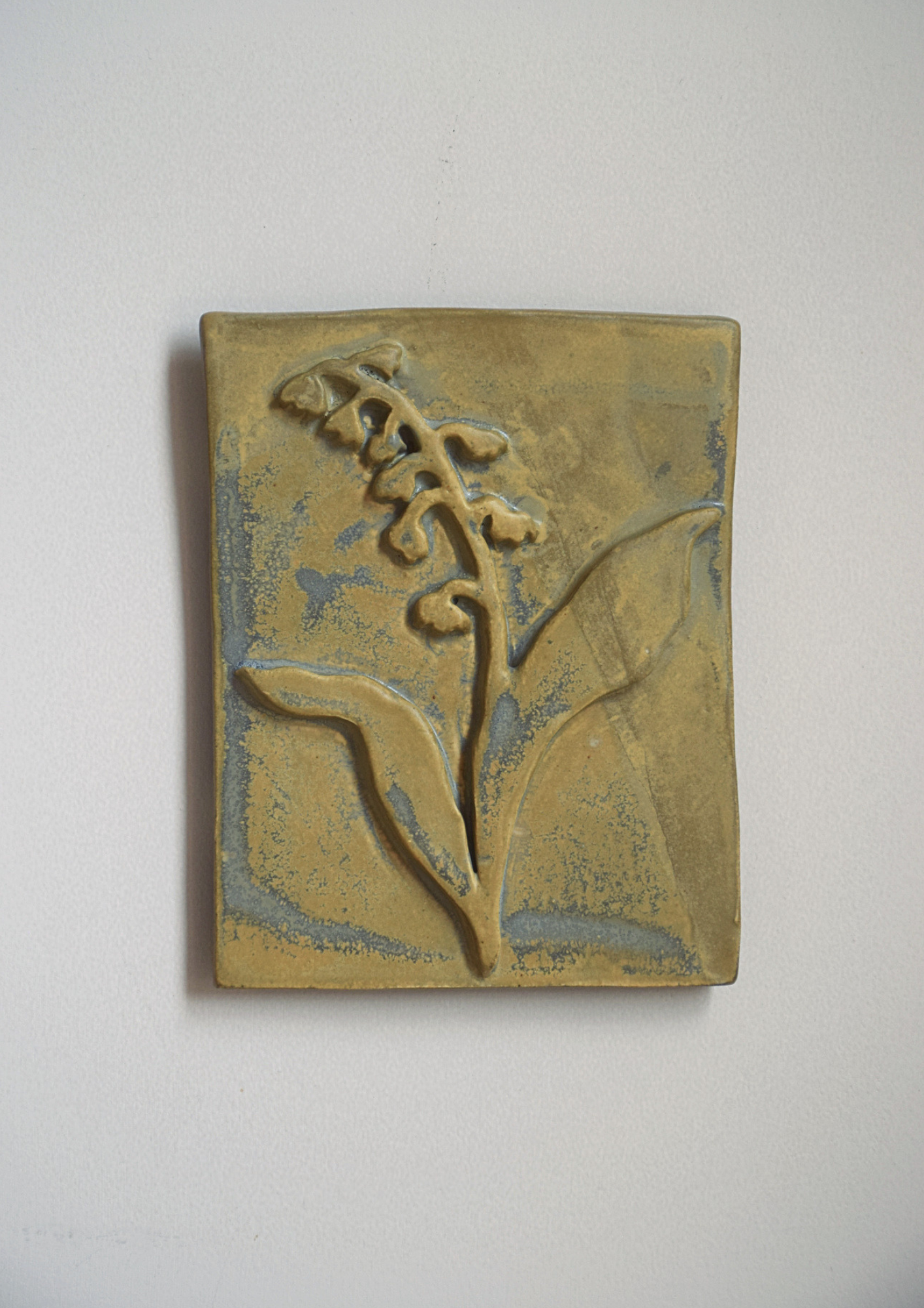 Une plaque en céramique avec une décoration en relief représentant une plante avec des feuilles et des petites fleurs dans un style artisanal, de couleur beige et légèrement bleutée.