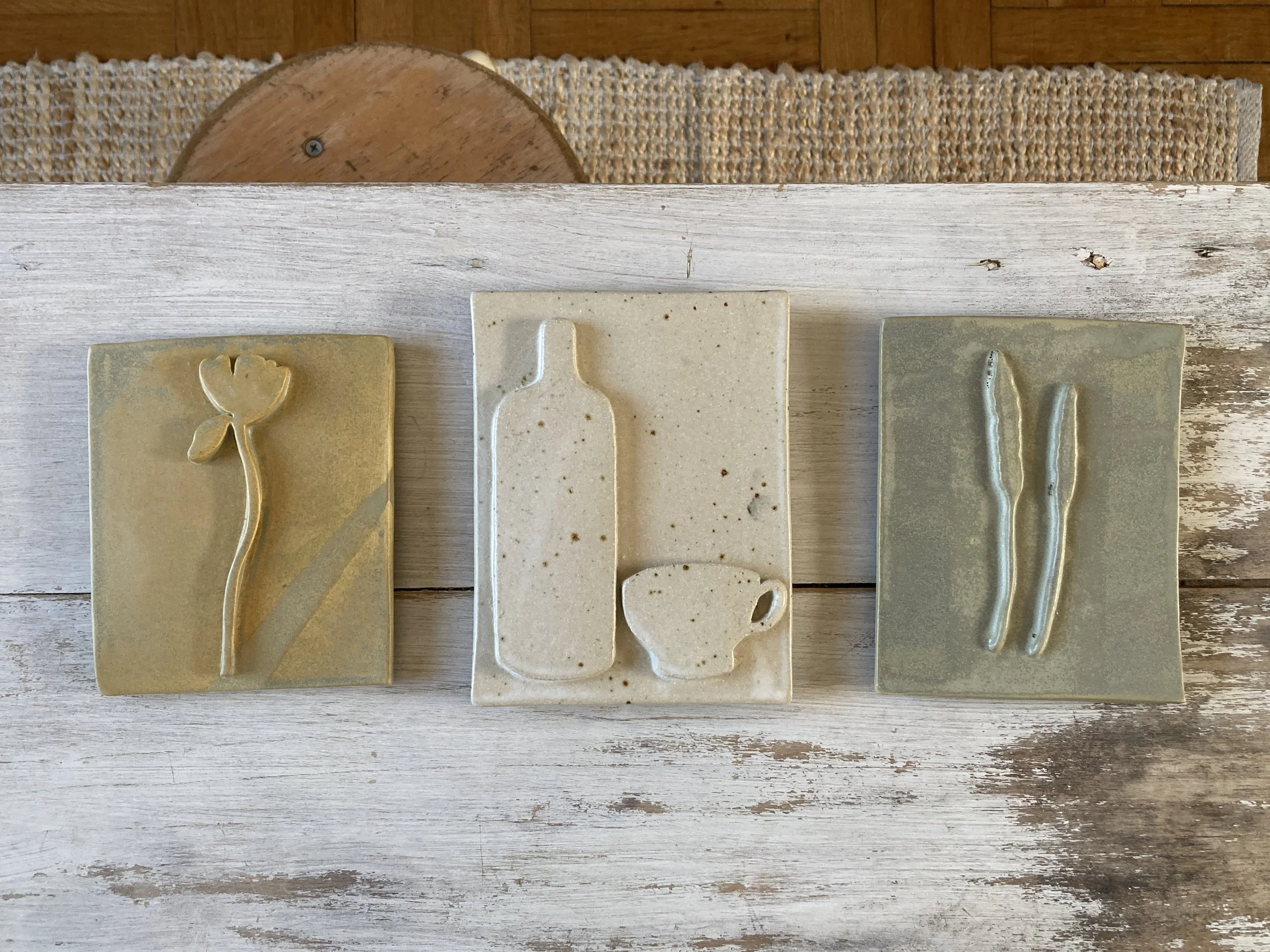 Trois  en céramique avec des formes de décoration mates sur un fond beige des trois côtés, posées sur une surface en bois. La plaque au centre a une forme de bouteille, tandis que les autres ont des formes abstraites. Le fond de l'image montre un tapis en laine et un mur en bois.