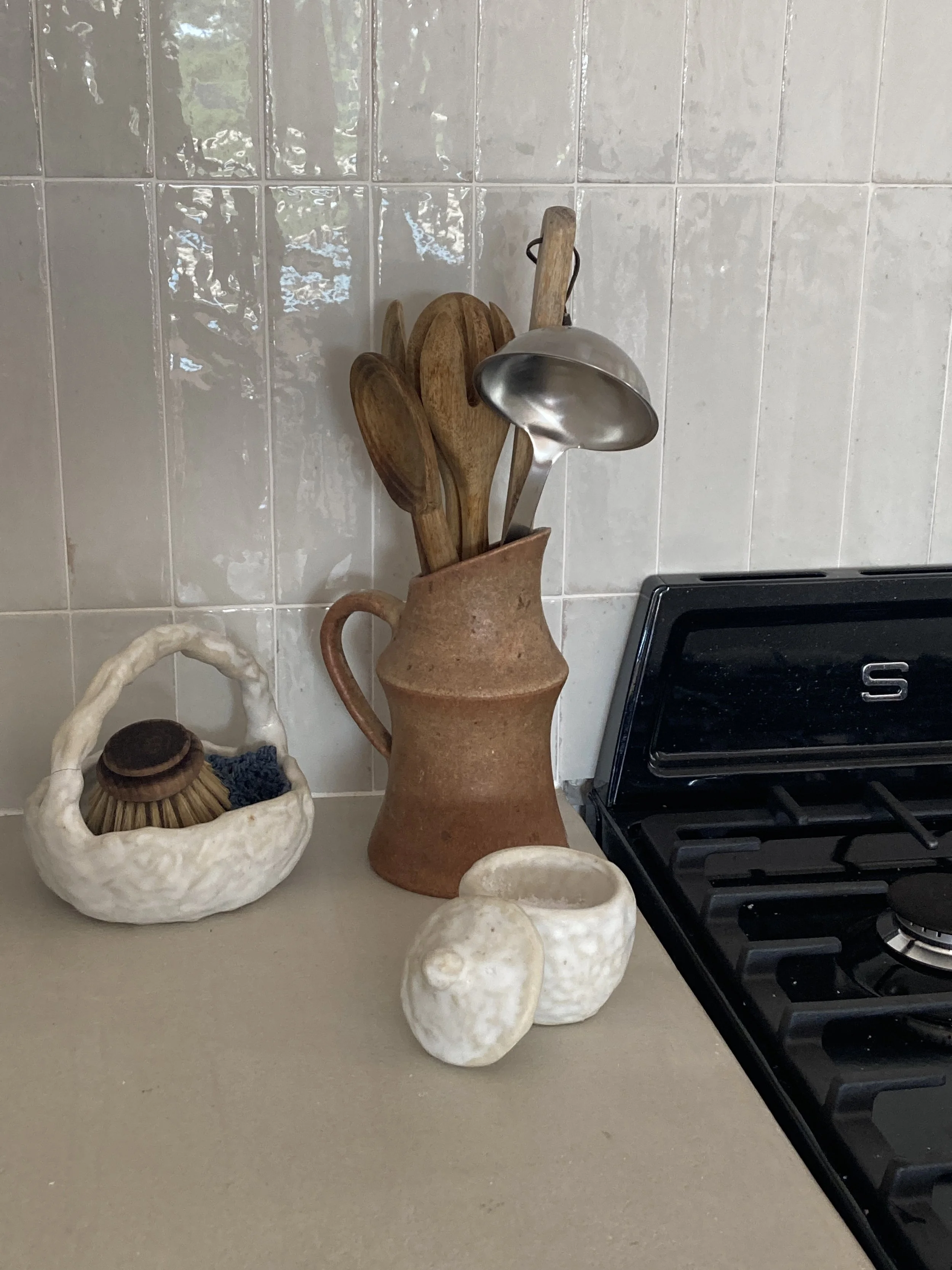 Une vase en terre cuite contenant des cuillères en bois et une louche en métal, située à côté d'une cuisinière au gaz avec un plan de cuisson noir, dans une cuisine avec un mur en carrelage clair.