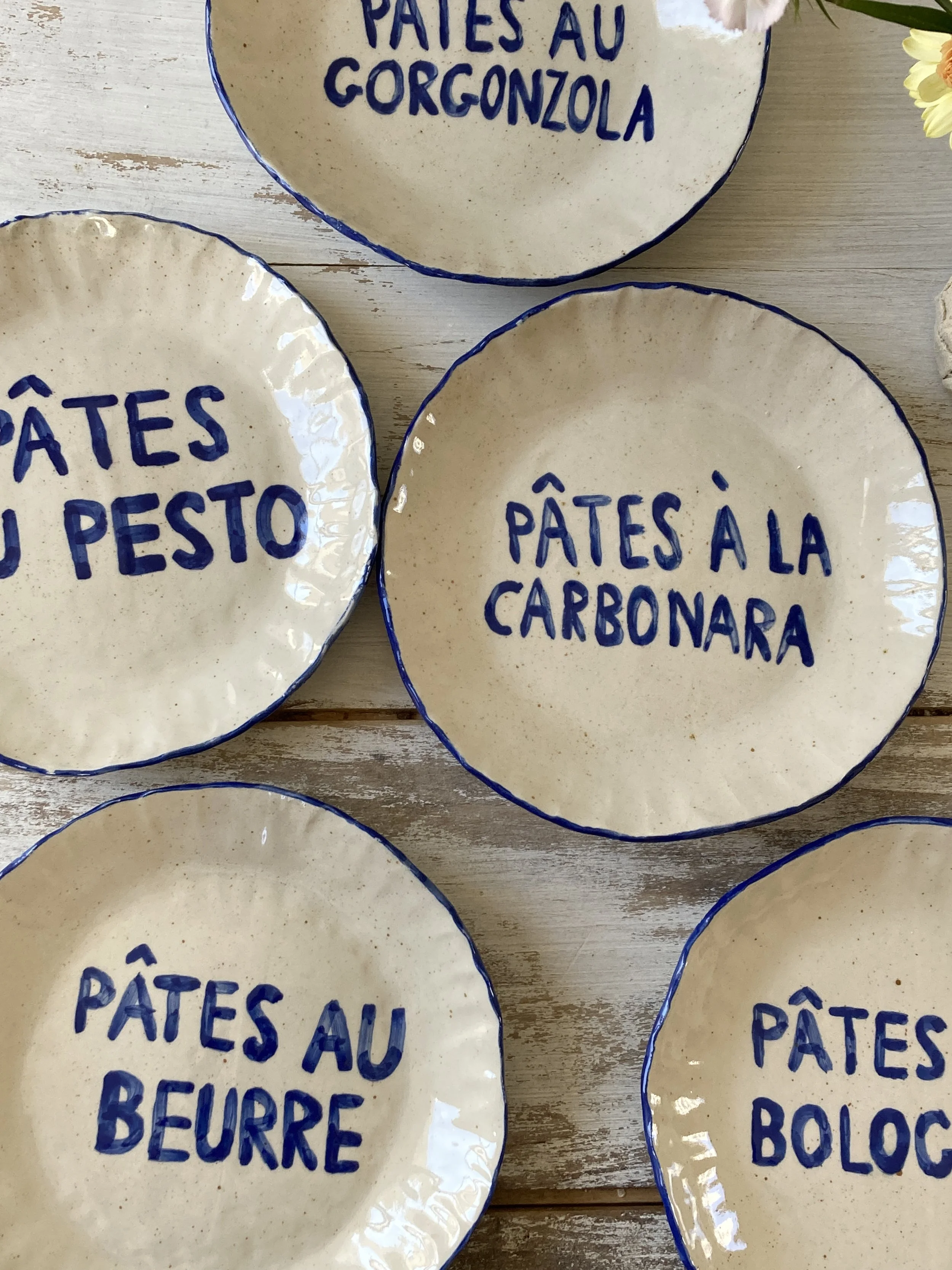 Assiettes en céramique avec inscriptions en bleu, indiquant différents types de pâtes, posées sur une table en bois. Certaines assiettes portent les inscriptions : « Pâtes au gorgonzola », « Pâtes à la carbonara » et « Pâtes al beurre ».