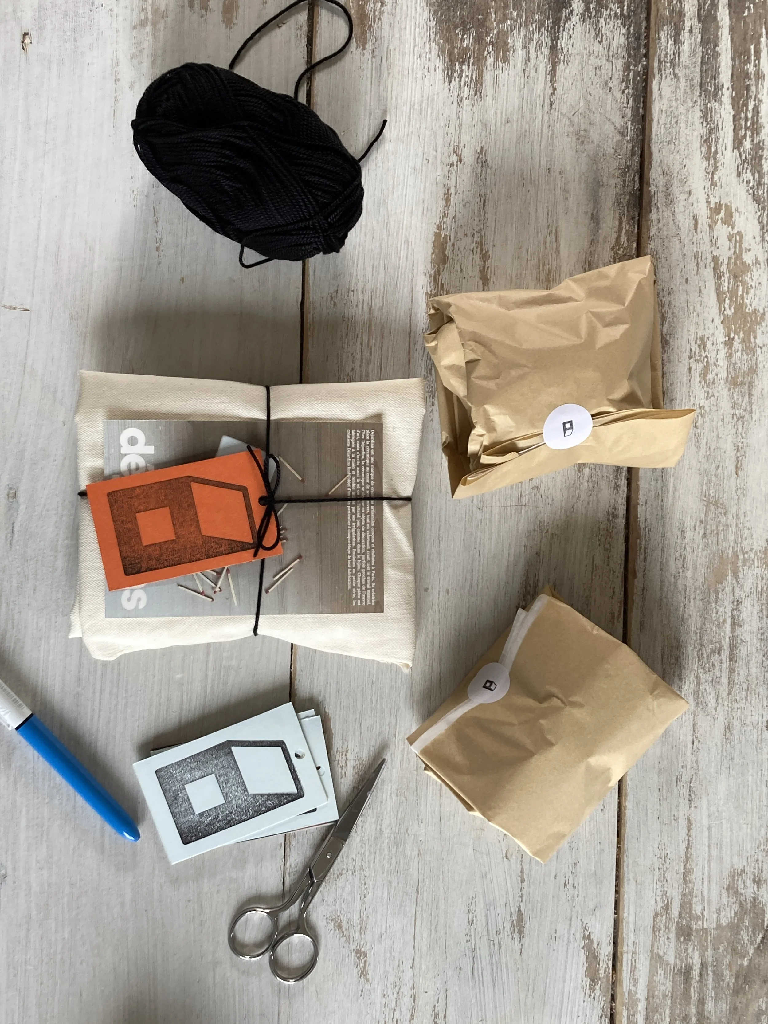 Divers objets pour un projet créatif : une boule de fil noir, plusieurs sachets en papier kraft avec des étiquettes, un stylo bleu, des ciseaux en acier inoxydable, des pinces, et des cartes avec des motifs en forme de maison en noir et blanc.