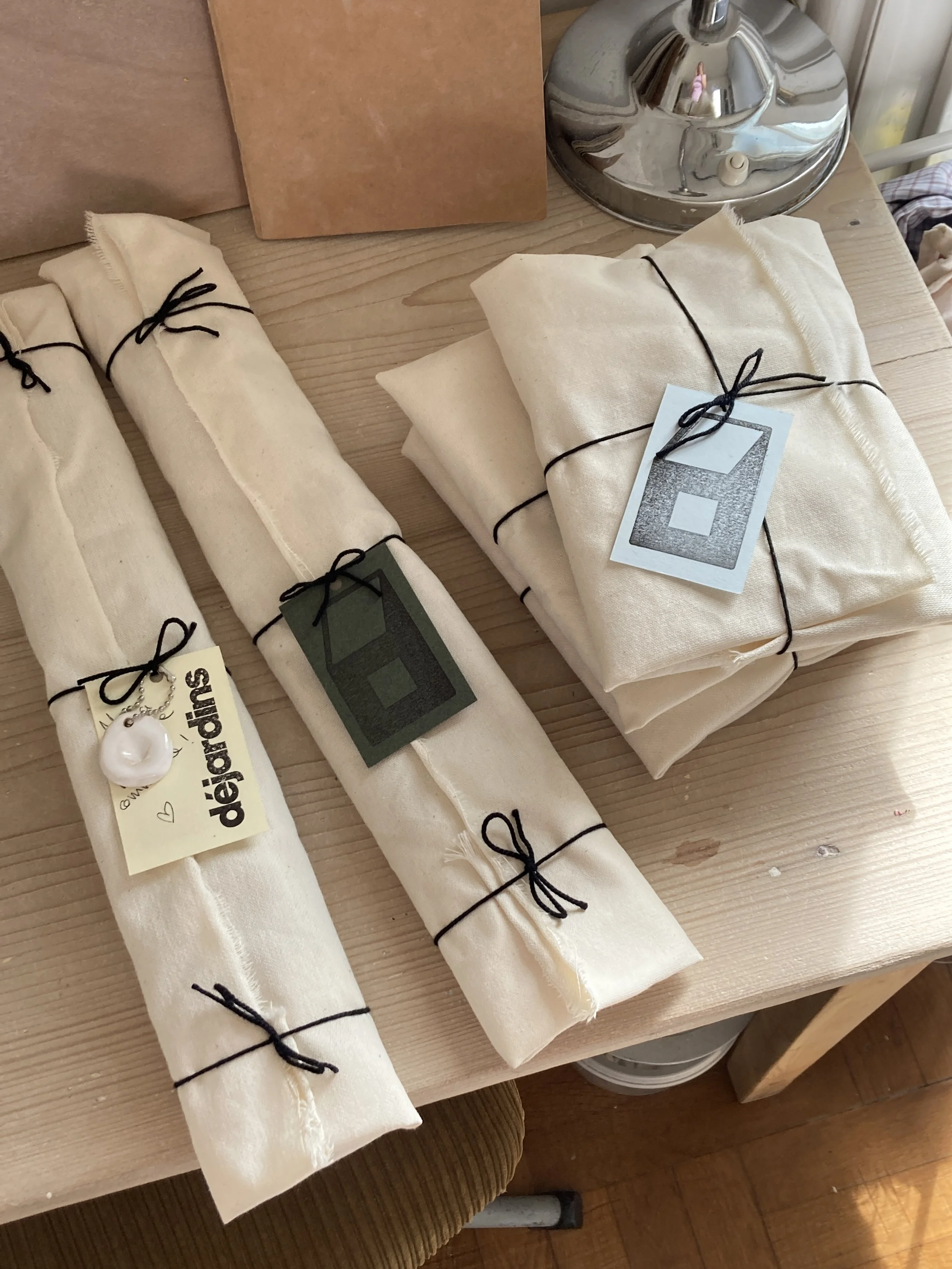 Plusieurs bouts de tissu blanc pliés et attachés avec des rubans noirs, posés sur une table en bois, avec des étiquettes.