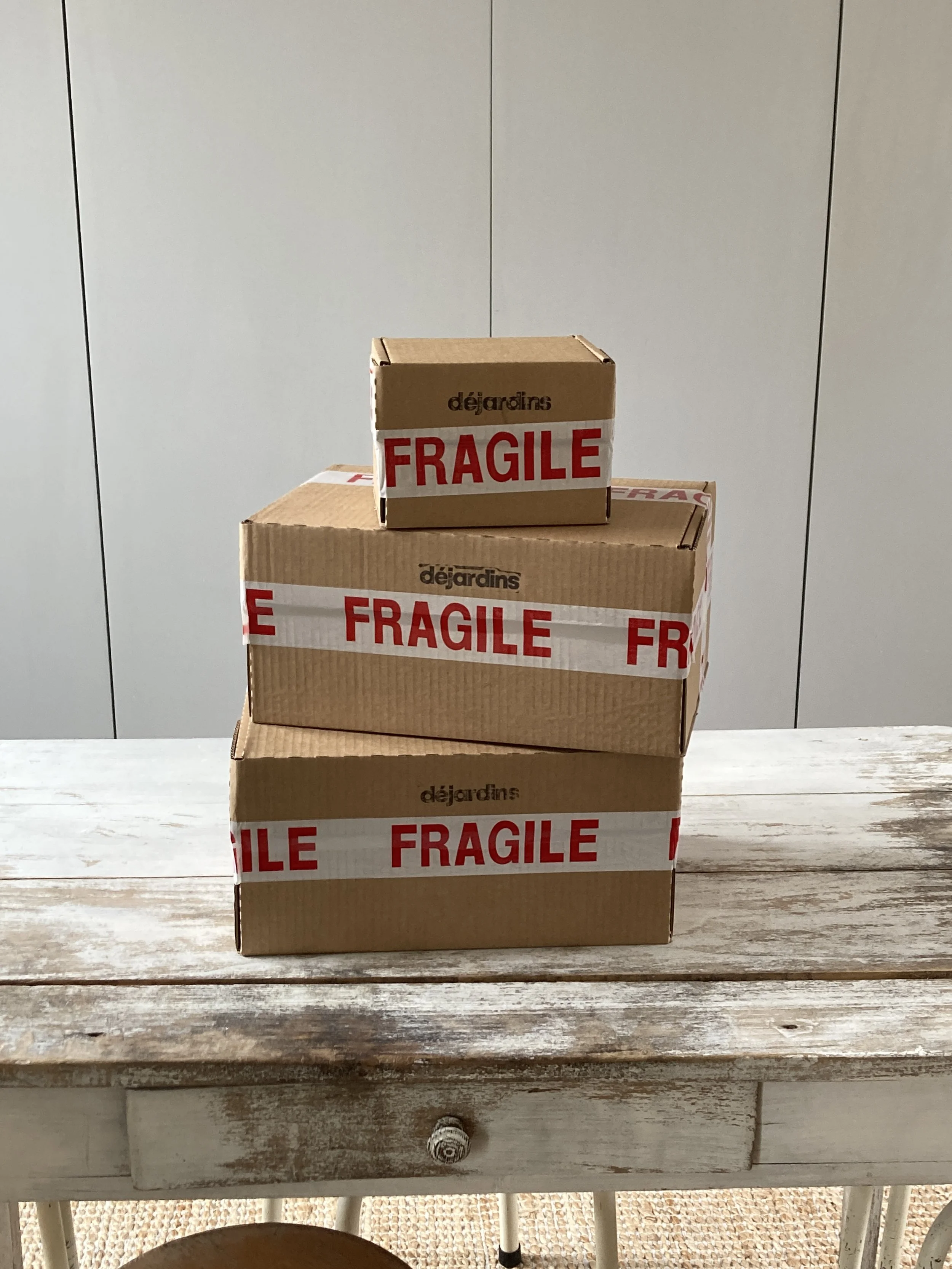 Trois cartons en carton marron avec des étiquettes "FRAGILE" en rouge, empilés sur une table en bois.