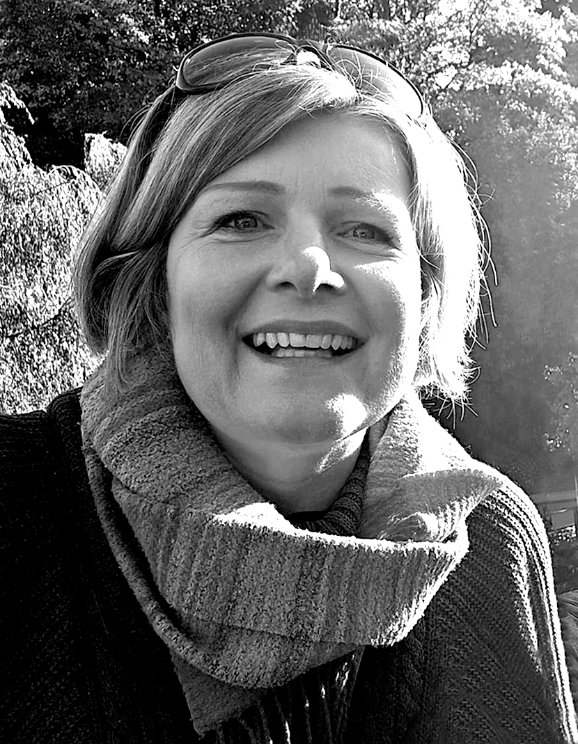 A woman smiling outdoors in black and white, wearing glasses on her head and a thick scarf around her neck.