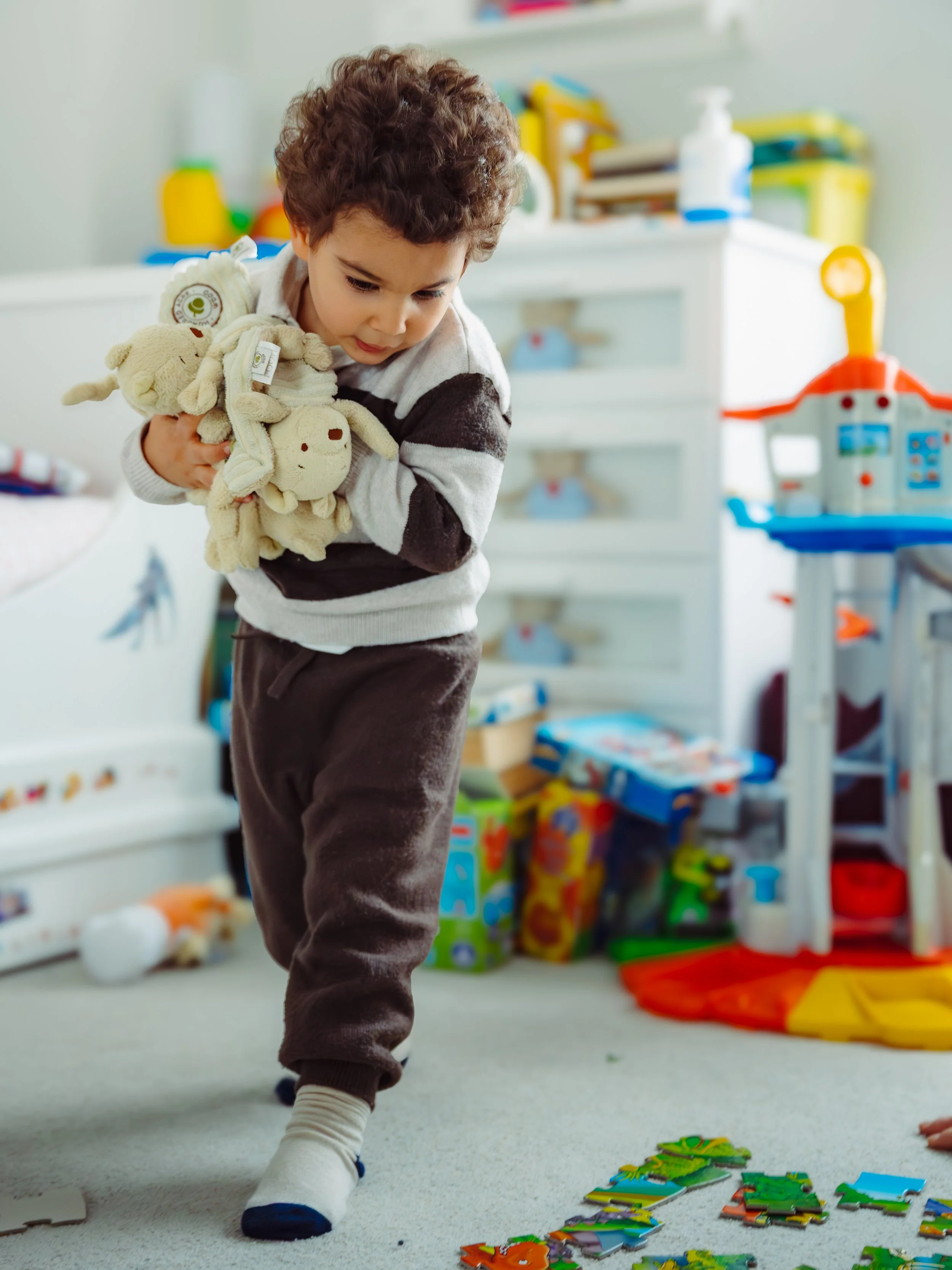 A young boy with curly hair and a striped sweater holding stuffed animal toys while standing in a playroom with toys and puzzles on the floor and shelves in the background.