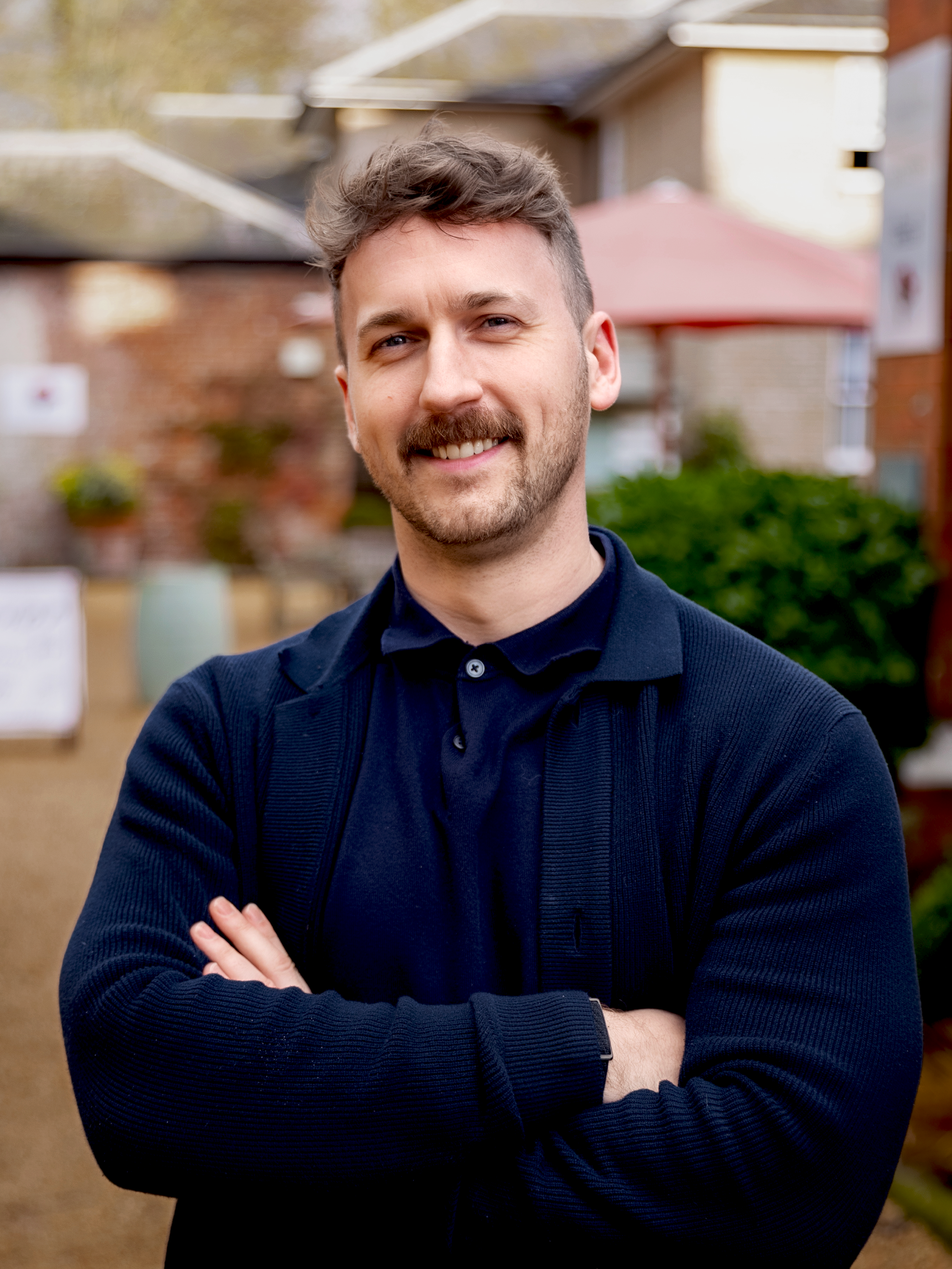 A smiling man with crossed arms, wearing a navy blue sweater, standing outdoors in front of blurred buildings and greenery.