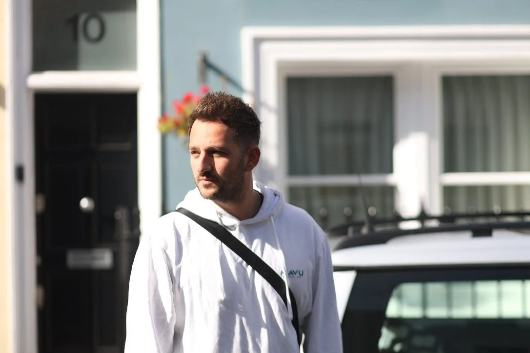 A man with short dark hair and a beard, wearing a white hoodie with a logo, standing outside in front of a house with a black door and a parked car, during daytime.