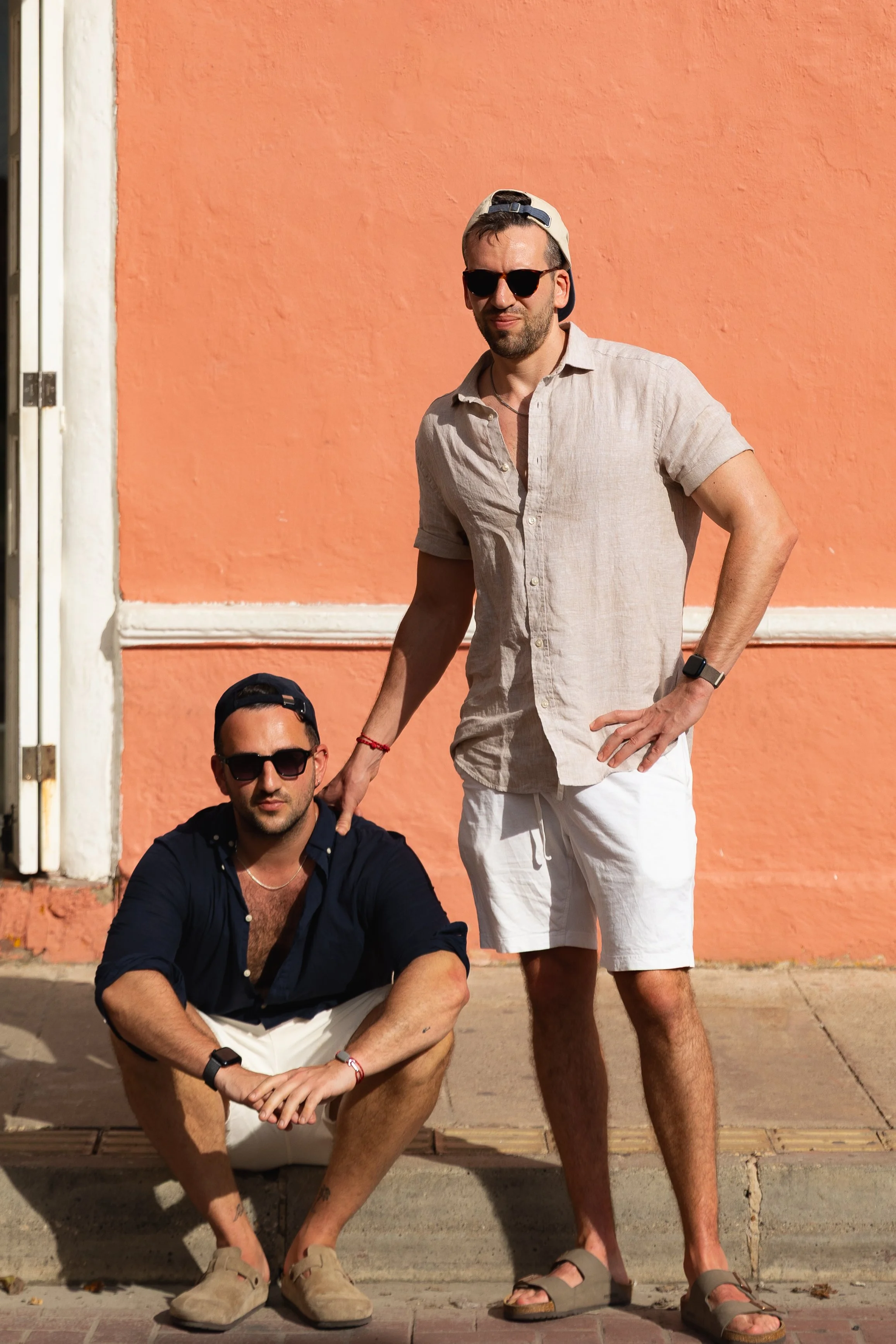 Two men pose in front of a pink wall, wearing sunglasses, casual summer clothes, and sandals, one squatting and one standing with his hand on the other's shoulder.