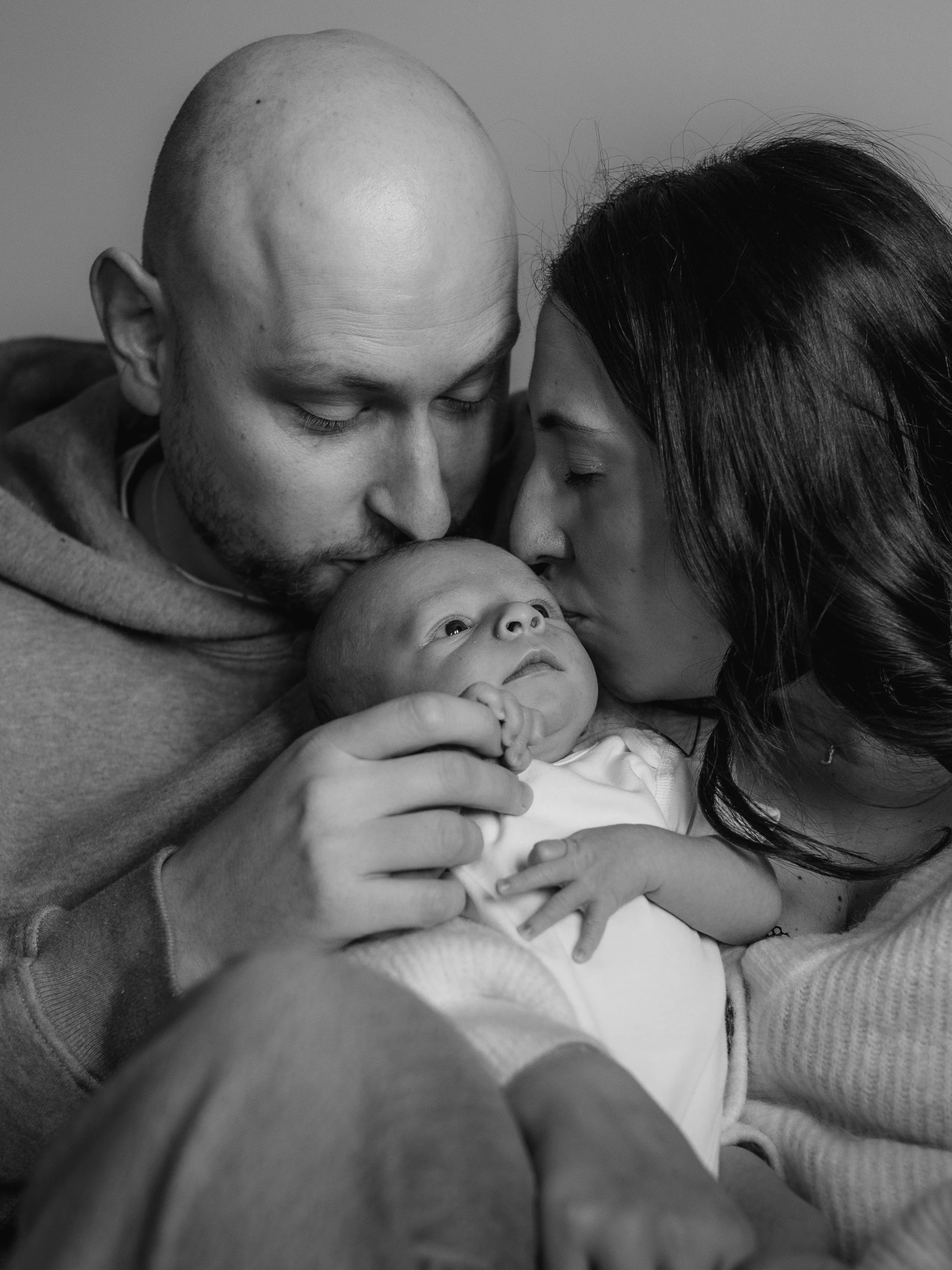 A family of three, including a man, woman, and baby, sharing a close embrace with the parents kissing the baby's head and face in black and white.