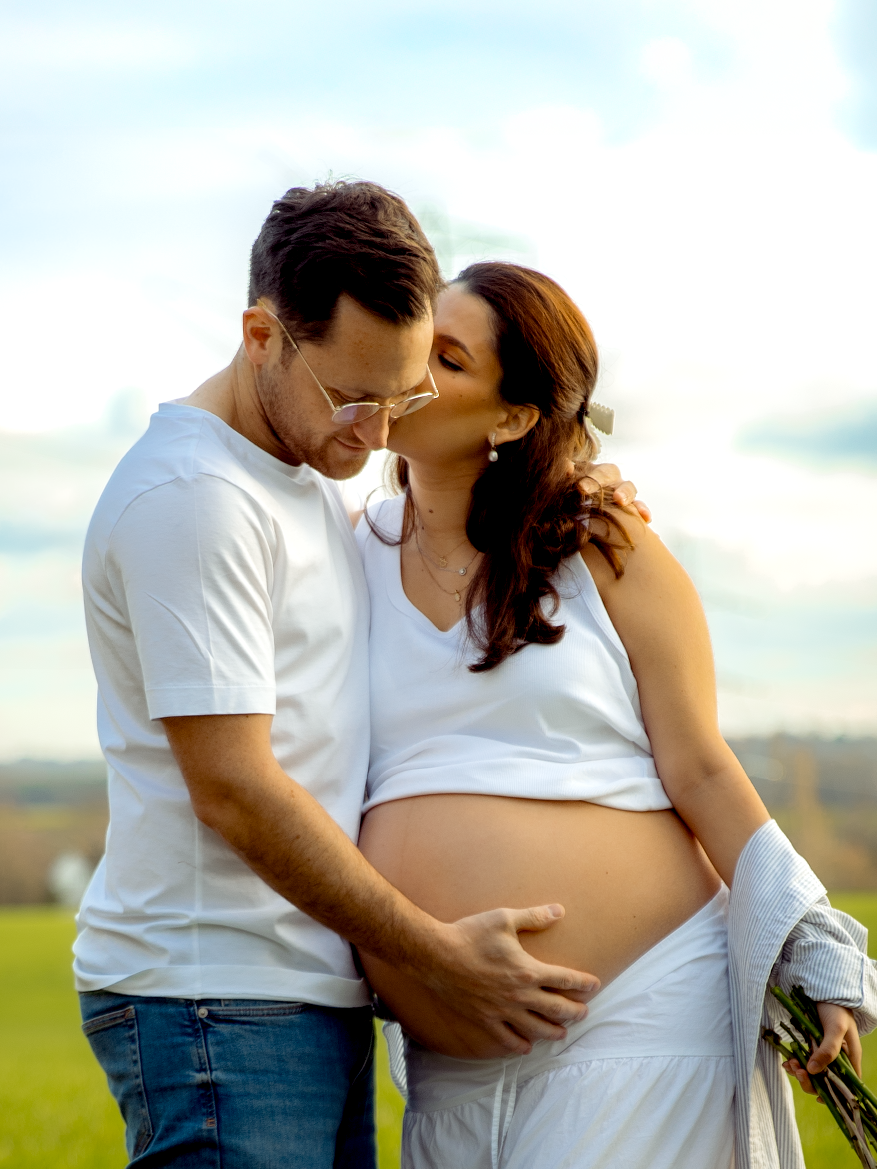 A pregnant woman with long brown hair and pearl earrings, wearing a white tank top, is kissing a man on the forehead while he gently touches her pregnant belly. The man has short brown hair, wears glasses, a white t-shirt, and jeans. They are outdoor