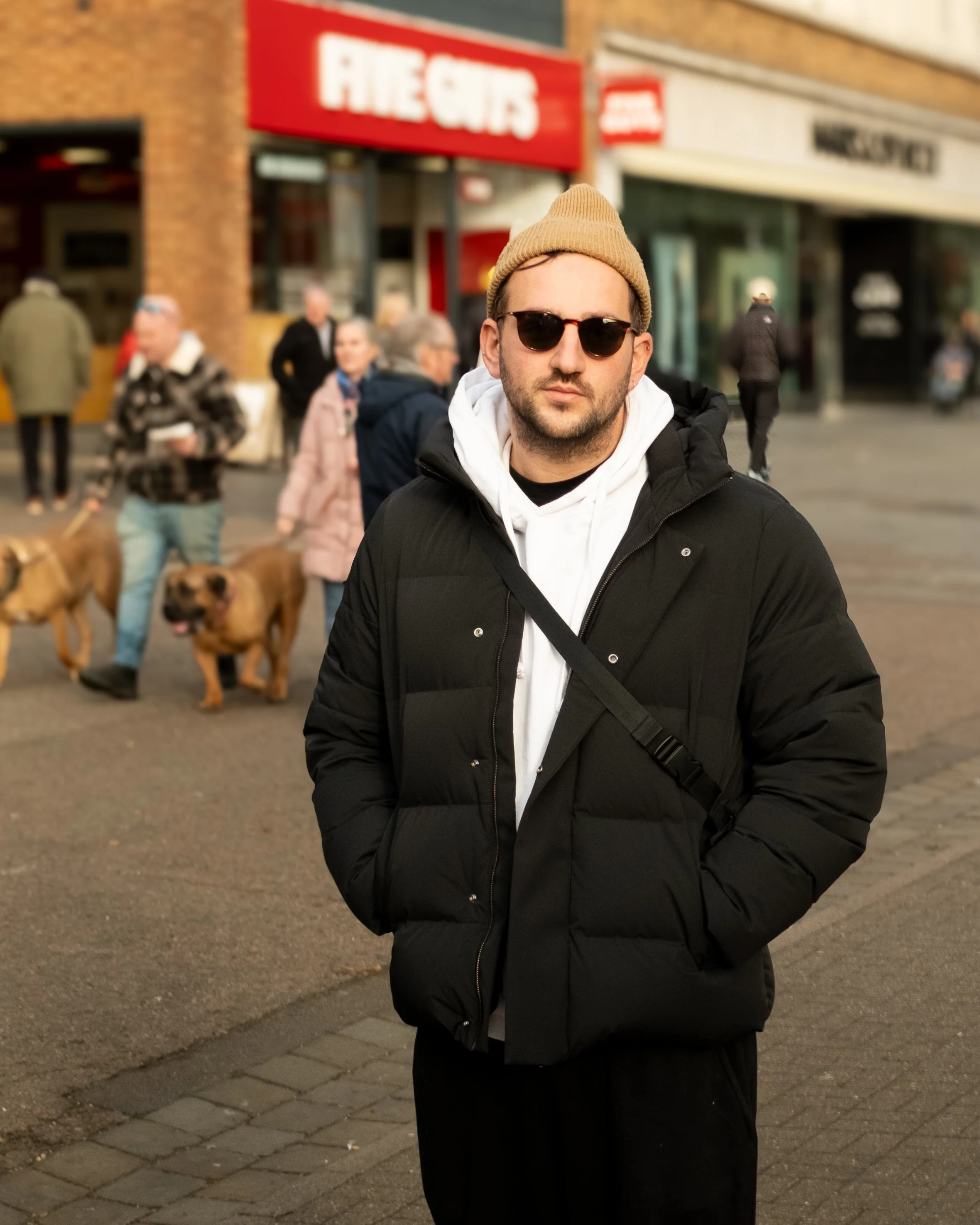 A man wearing sunglasses, a tan beanie, and a black puffer jacket standing on a city sidewalk with people and dogs in the background.