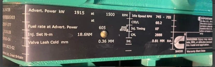 Close-up of a metal nameplate on machinery displaying various technical specifications such as power in kW, fuel rate, injection settings, and valve lash. Also includes idle speed RPM and identification numbers.