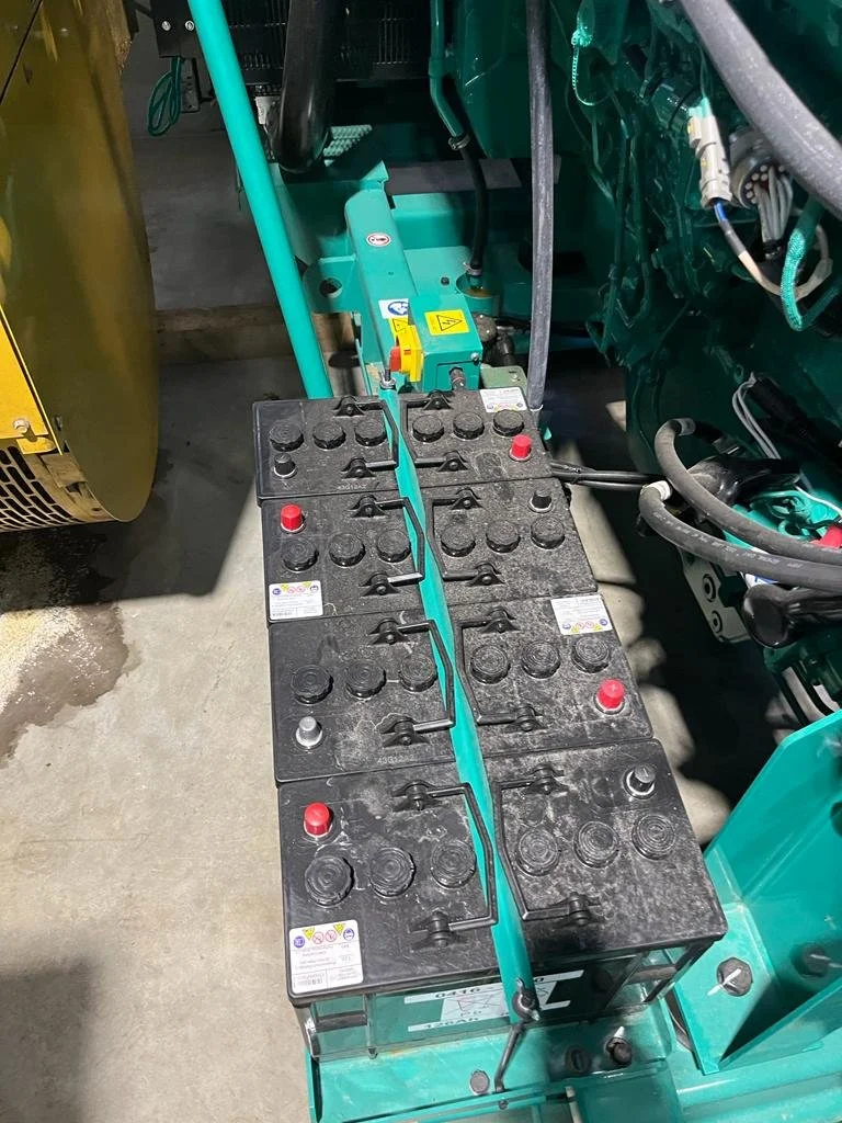 Close-up of industrial batteries on a metal frame, connected by cables, with machinery components nearby.