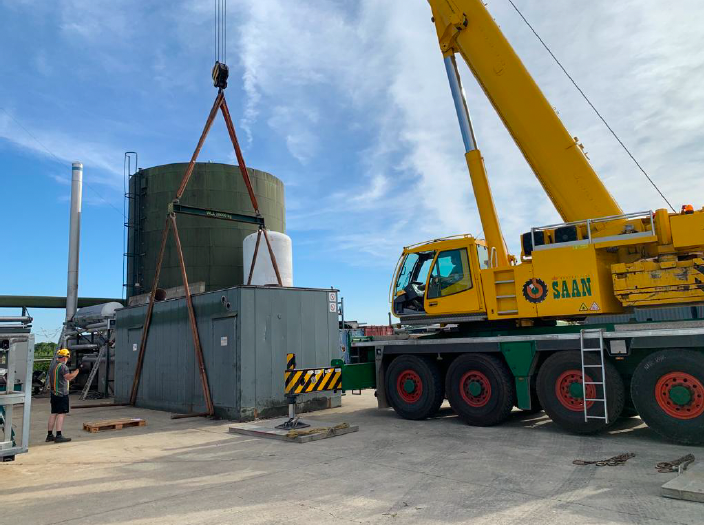 Construction site featuring a yellow crane lifting a large metal container beside industrial tanks and machinery, with a worker observing.