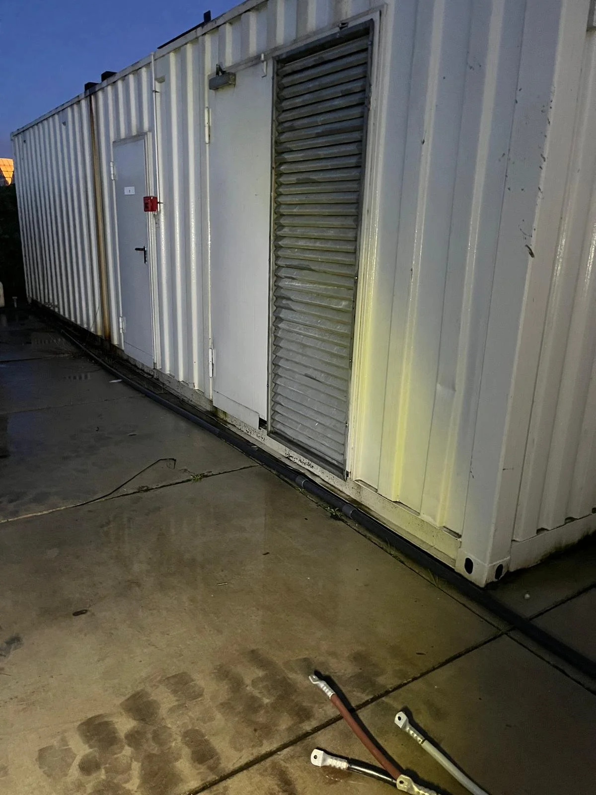 A metal storage container with doors and vents, resting on a concrete surface with some cables on the ground in front of it.