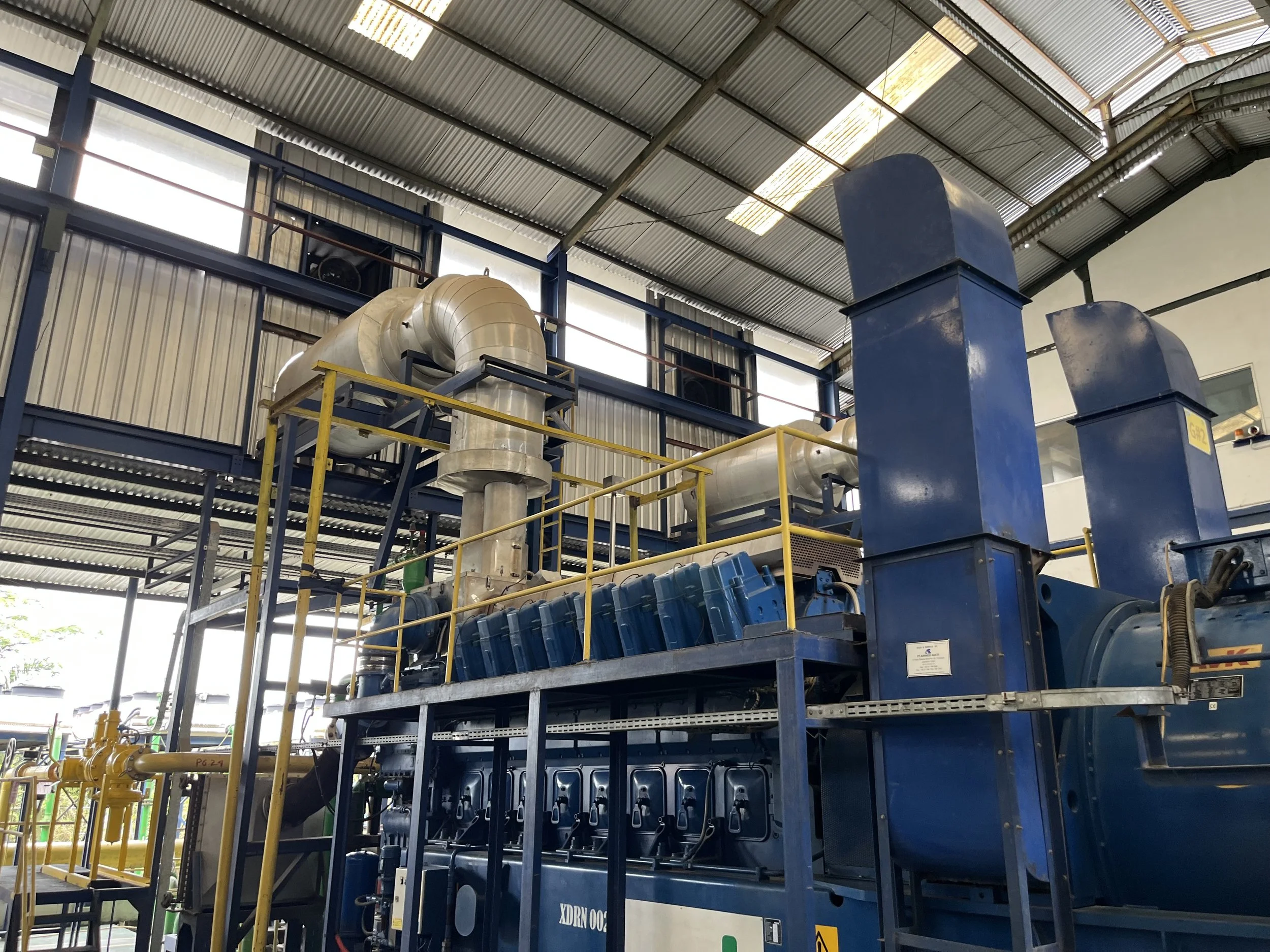 Industrial machinery inside a factory with large blue metal structures, yellow railings, and air ducts under a corrugated metal roof.
