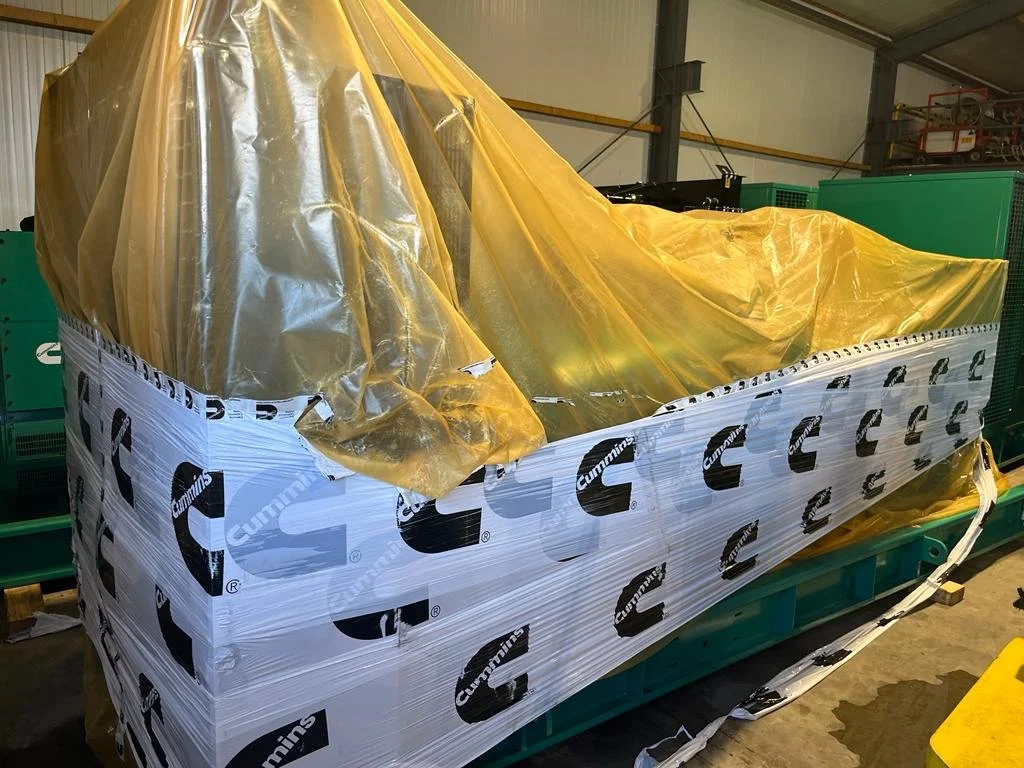 Large machine wrapped in Cummins branded plastic covering and yellow tarp in an industrial setting.