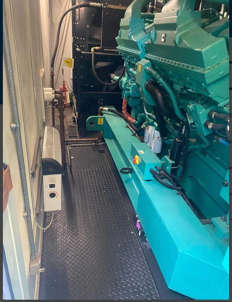 Large industrial diesel generator in an enclosed space, featuring a green engine and metal components. The floor has a diamond plate pattern.