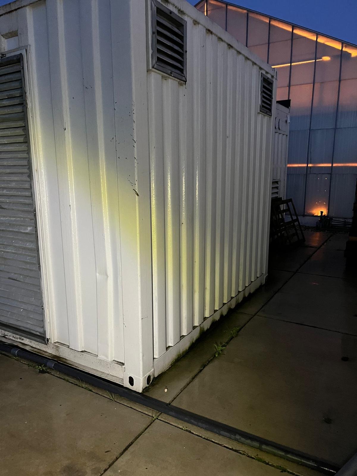 White shipping container with vents, located outdoors near a modern greenhouse, at dusk.