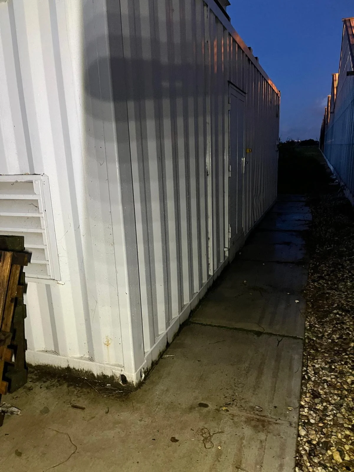 Metal shipping container with a door on a paved and gravel-covered area during evening.