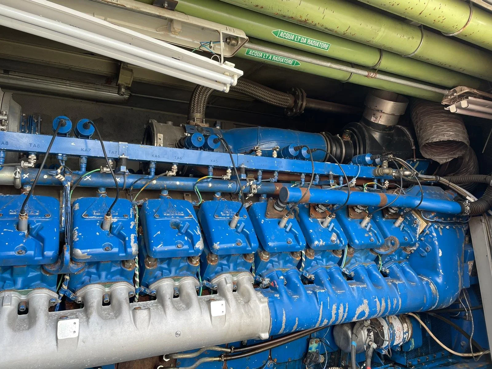 Close-up of a large industrial blue engine with multiple cylinders, pipes, and wires. Green pipes are visible above the engine, labeled with text. The engine appears heavily used and painted in blue. Metal and mechanical components are prominently sh