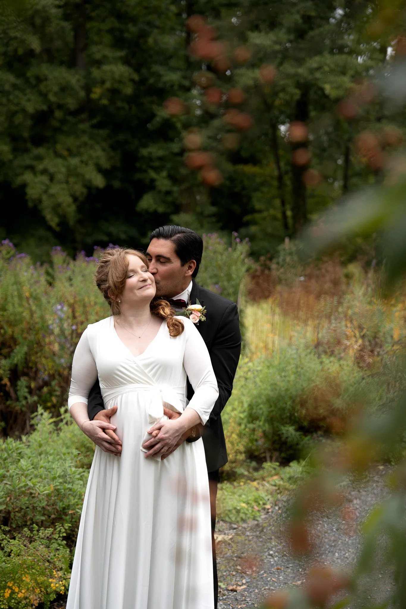 Ein Brautpaar steht in einem Garten, der Bräutigam küsst die schwangere Braut auf die Stirn, umgeben von grünen Pflanzen und Bäumen.