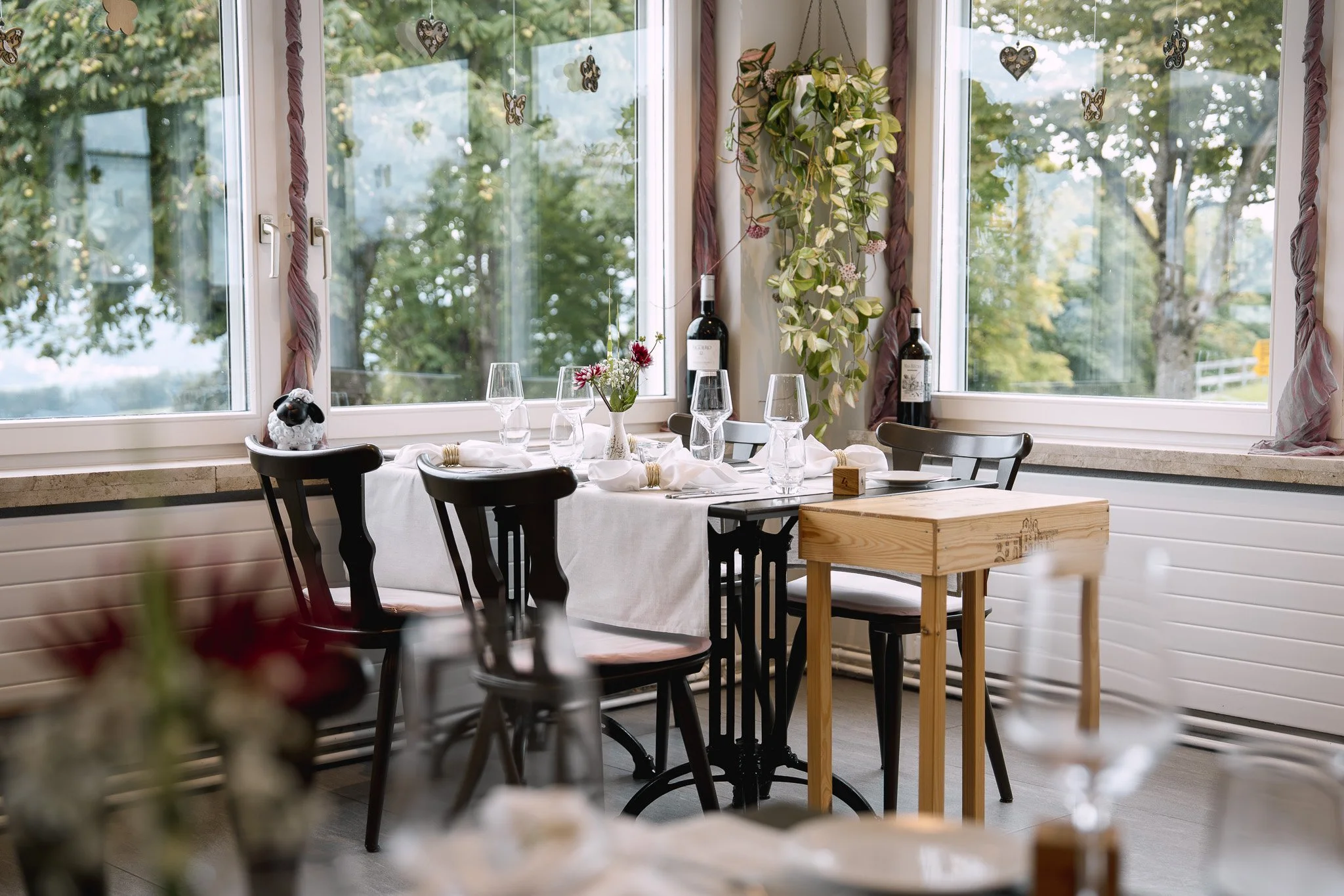 Ein gedeckter Esstisch mit Weingläsern, Blumen und Weinfässern vor einem Fenster mit grünen Bäumen im Hintergrund.