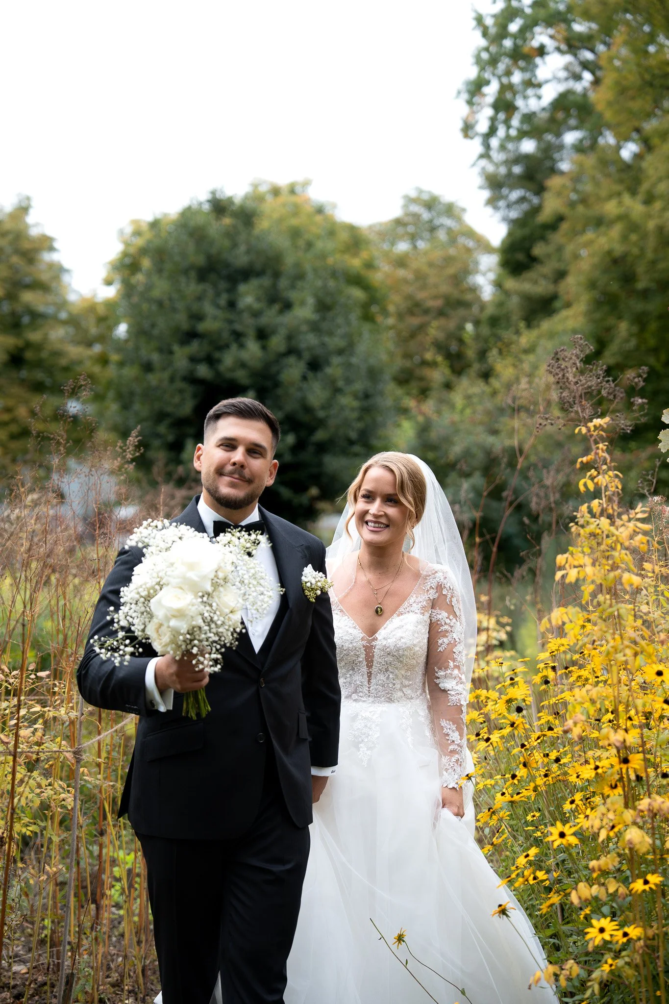Ein Brautpaar im Hochzeitsoutfit in einem Garten, umgeben von gelben Blumen und grünen Bäumen, beim Fotoshooting im Freien.