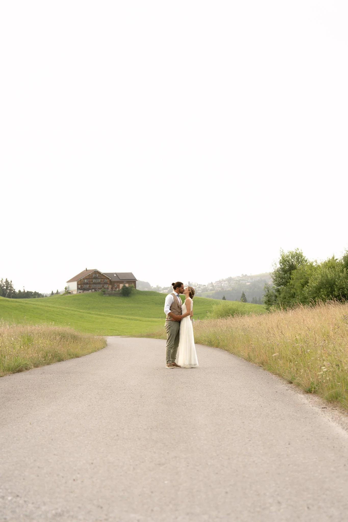 Ein Brautpaar küsst sich auf einer ländlichen Straße, umgeben von grünen Feldern und sanften Hügeln, mit einem Haus im Hintergrund.