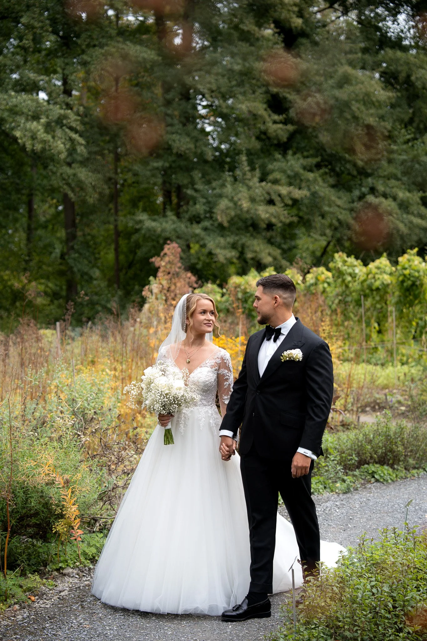 Ein Brautpaar steht im Freien, umgeben von Büschen und Bäumen, bei einer Hochzeit. Die Braut trägt ein weißes Kleid mit Spitze, eine Schleier und hält einen Blumenstrauß. Der Bräutigam in einem schwarzen Anzug mit Fliege hält ihre Hand und schaut sie