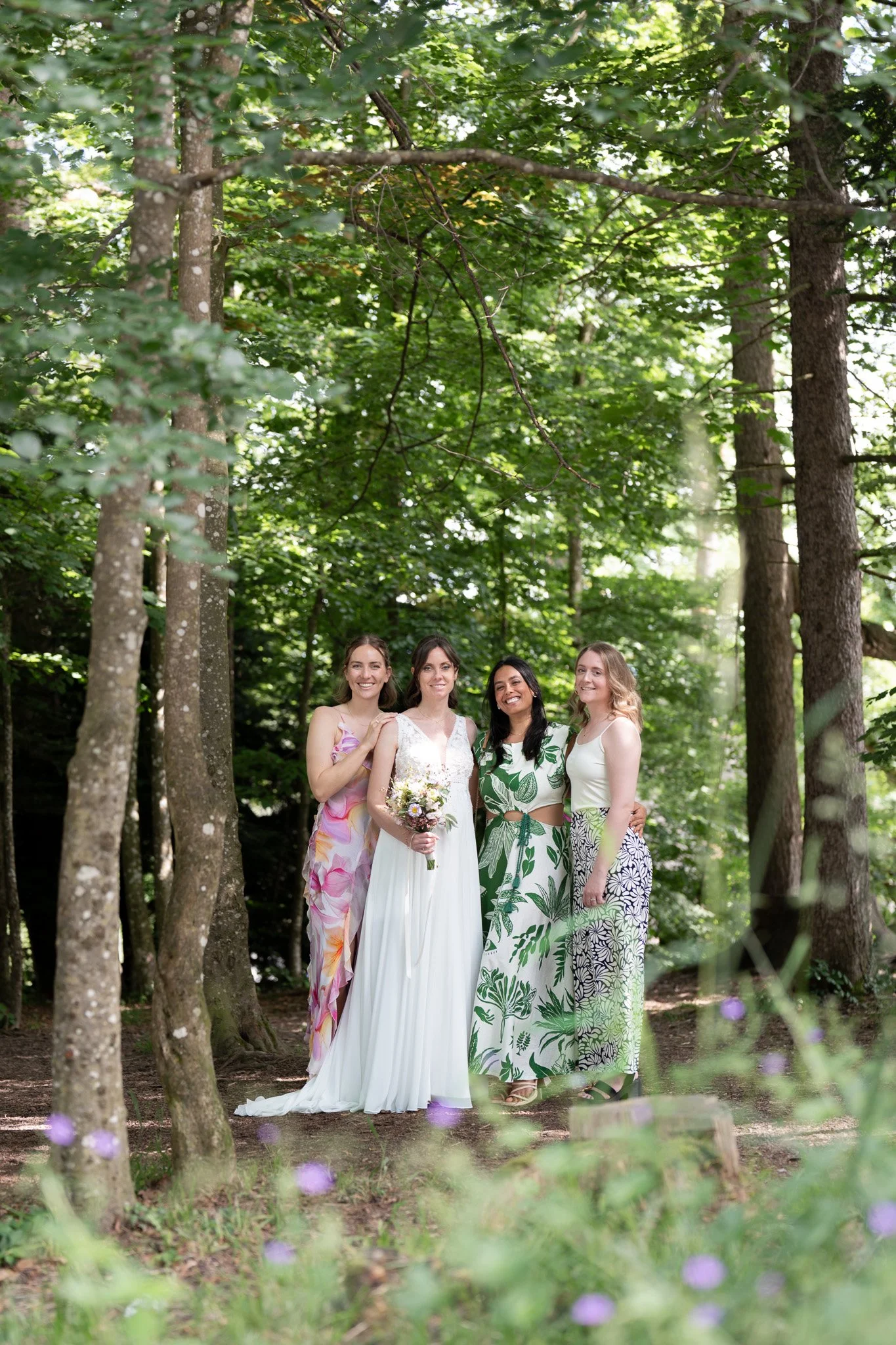 Vier Frauen stehen im Wald, eine trägt ein weißes Brautkleid mit Blumenstrauß, die anderen tragen bunte, sommerliche Kleider.