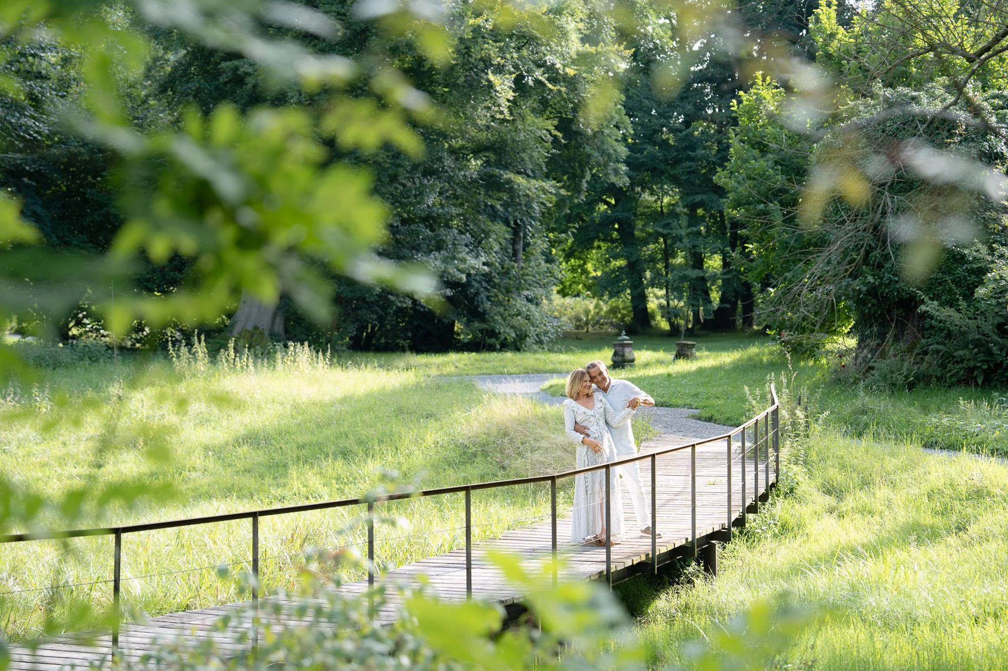 Ein Paar auf einer Holzbrücke in einem grünen Park bei Tageslicht, umgeben von Bäumen und Gras, beim Spaziergang.