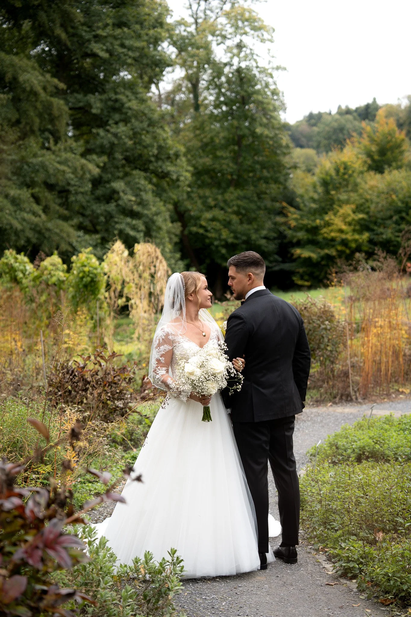 Braut und Bräutigam stehen sich in einem Garten bei einer Hochzeit, die Braut hält einen Blumenstrauß, die Umgebung ist grün mit Bäumen und Büschen im Herbst.