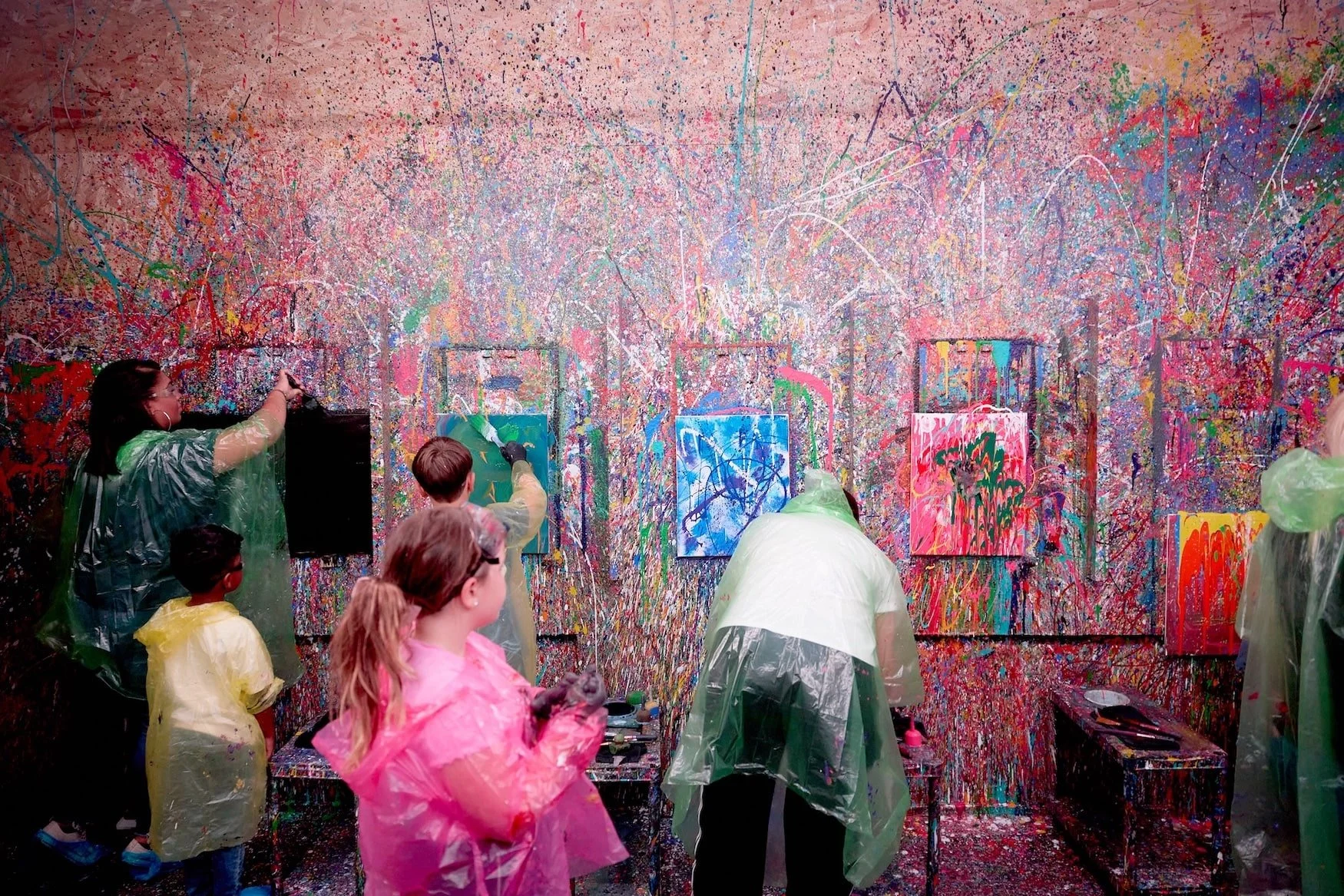 People wearing protective smocks creating abstract paintings on a colorful splattered wall in a paint and art studio.