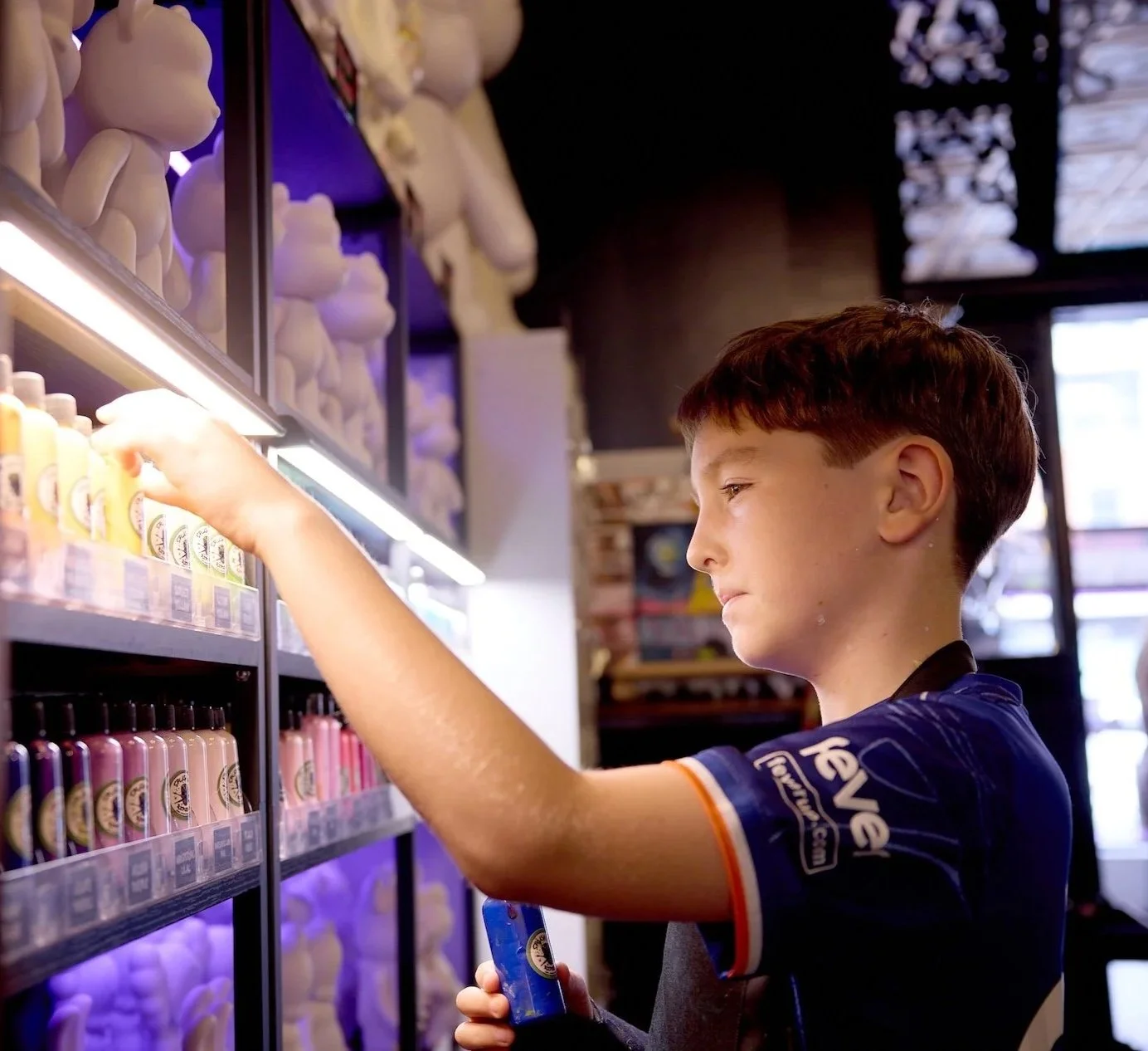 A young boy shopping at a store, reaching for a bottle on a shelf filled with various bottles and plush toys in the background.