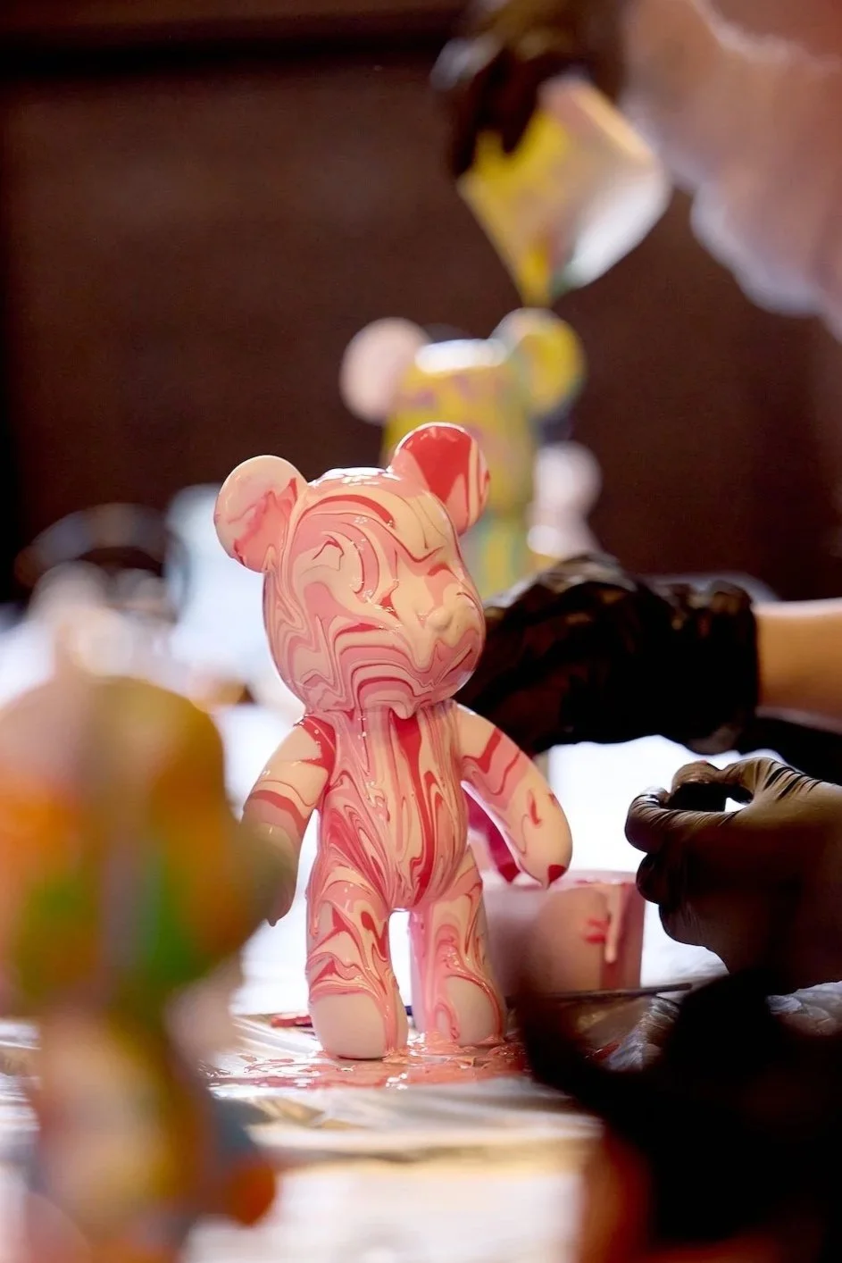 Marbled pink and white teddy bear sculpture being painted or decorated, with people wearing black gloves working on it.