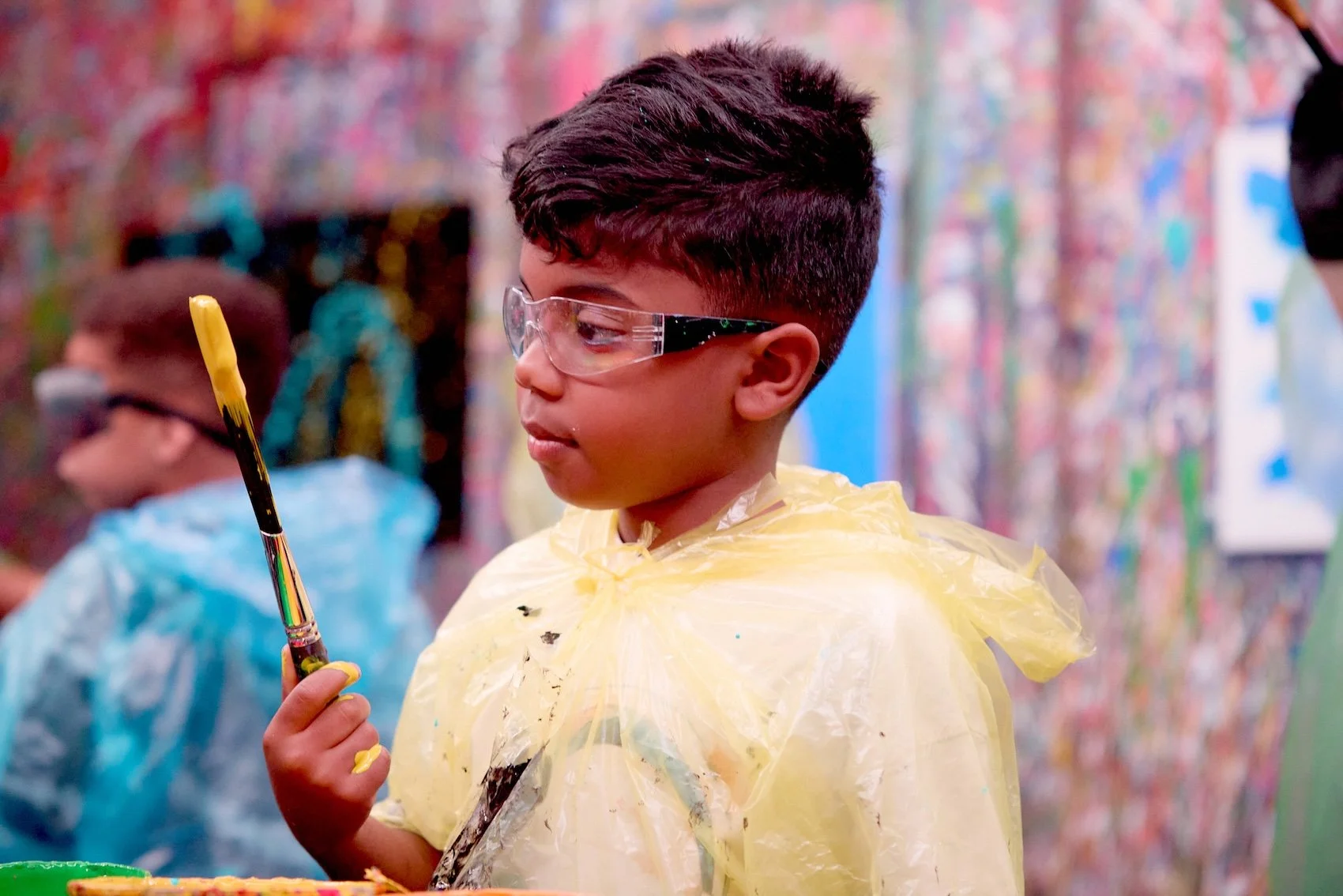 A young boy with glasses wearing a yellow plastic smock, holding a paintbrush with yellow paint, in an art studio with colorful abstract art on the walls.