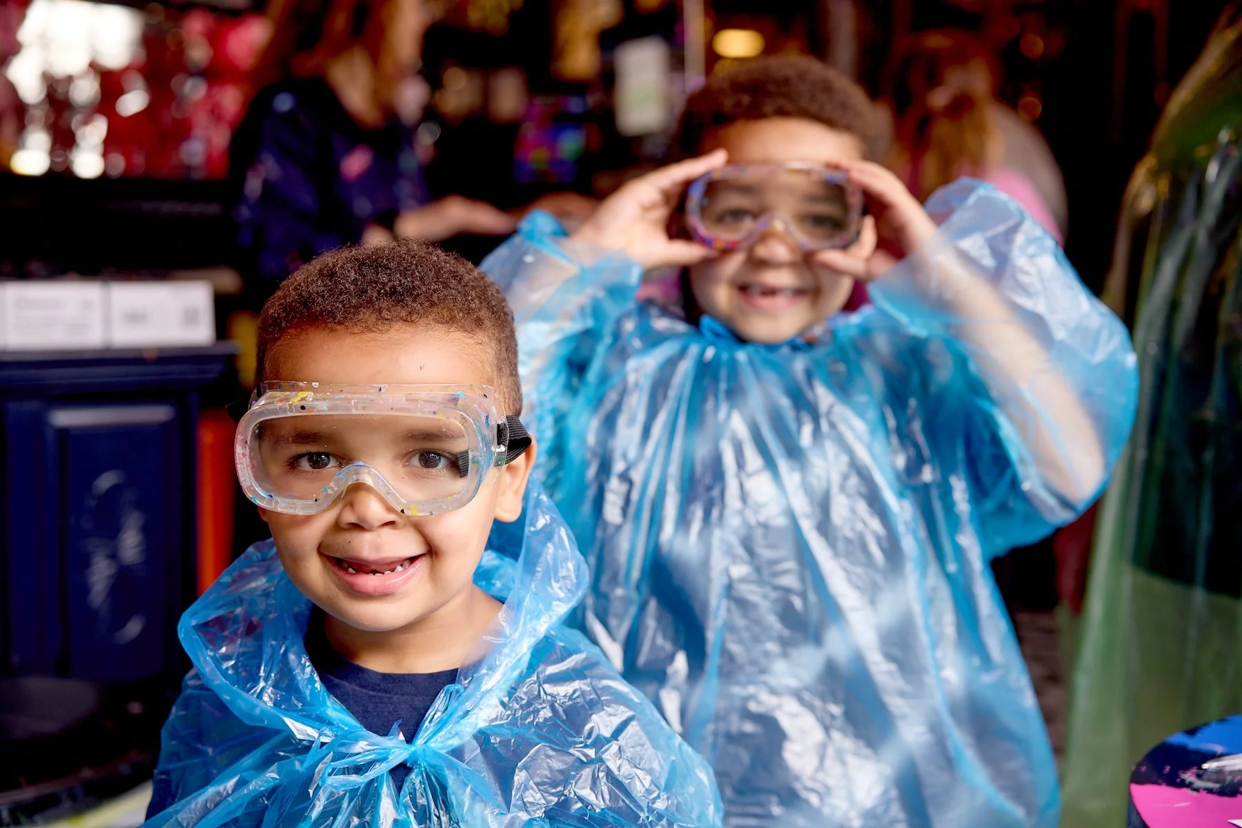 Two smiling young boys wearing safety goggles and blue plastic smocks, one holding goggles to his face, in a colorful indoor setting.