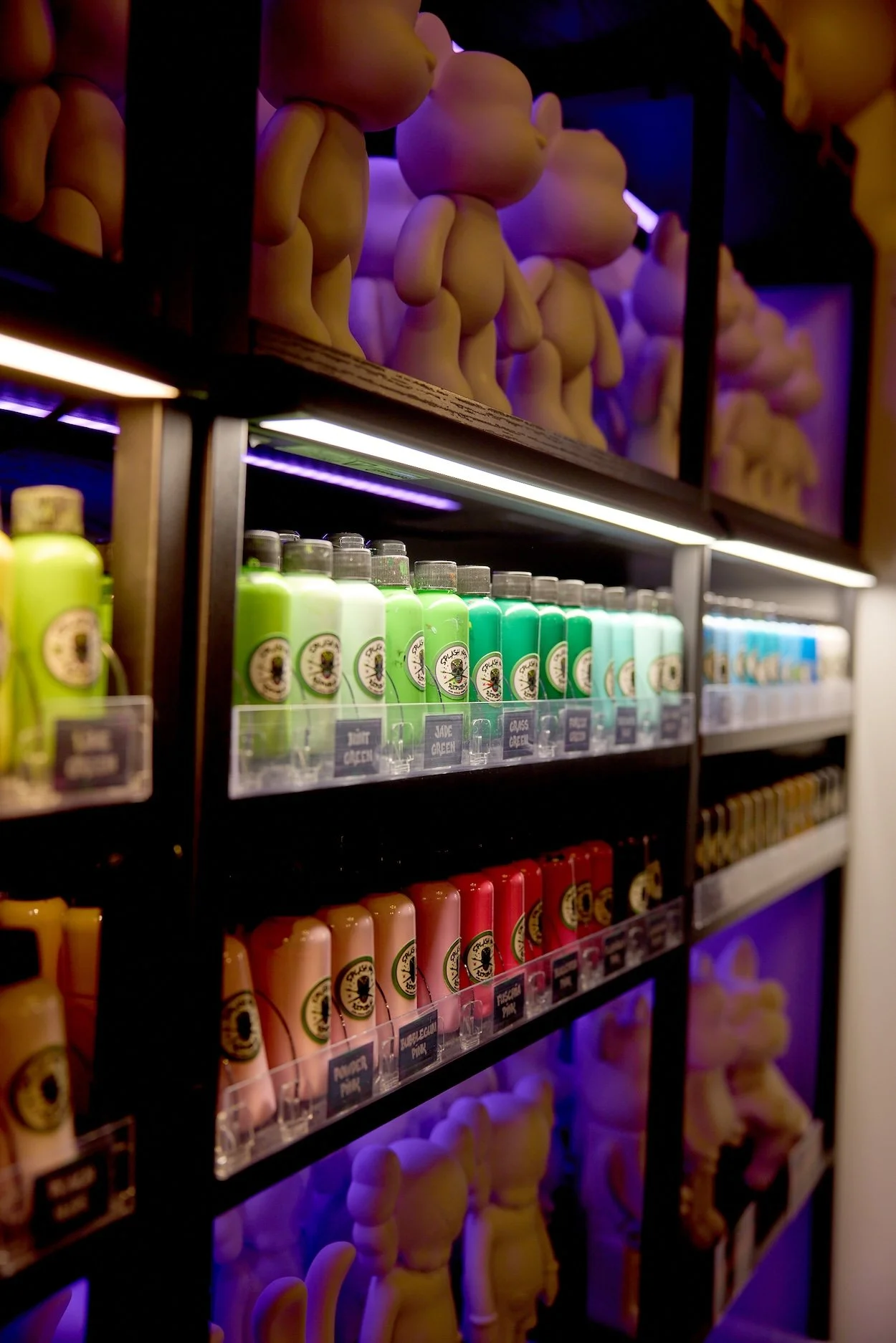 Display of colorful bottles on black shelving with teddy bear figurines on the top shelves, illuminated by purple and white lighting.