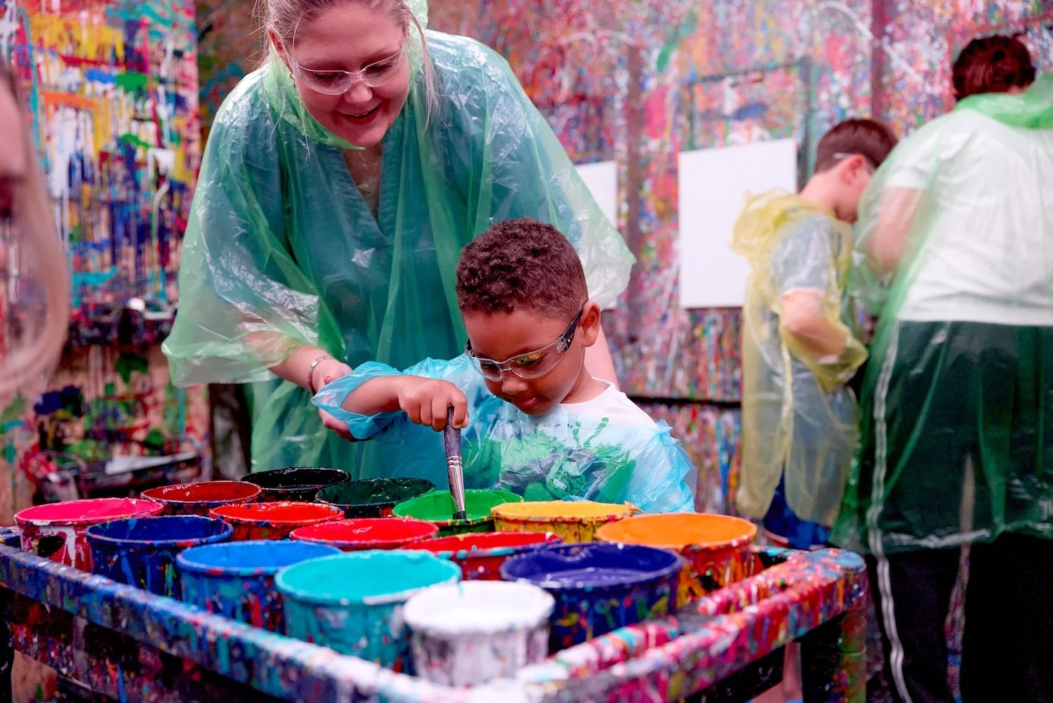 A woman and a young boy wearing protective glasses and plastic aprons are painting with bright colors in an abstract art studio. The girl is helping and guiding the boy as he paints with a brush. The background has a colorful, splattered paint wall.