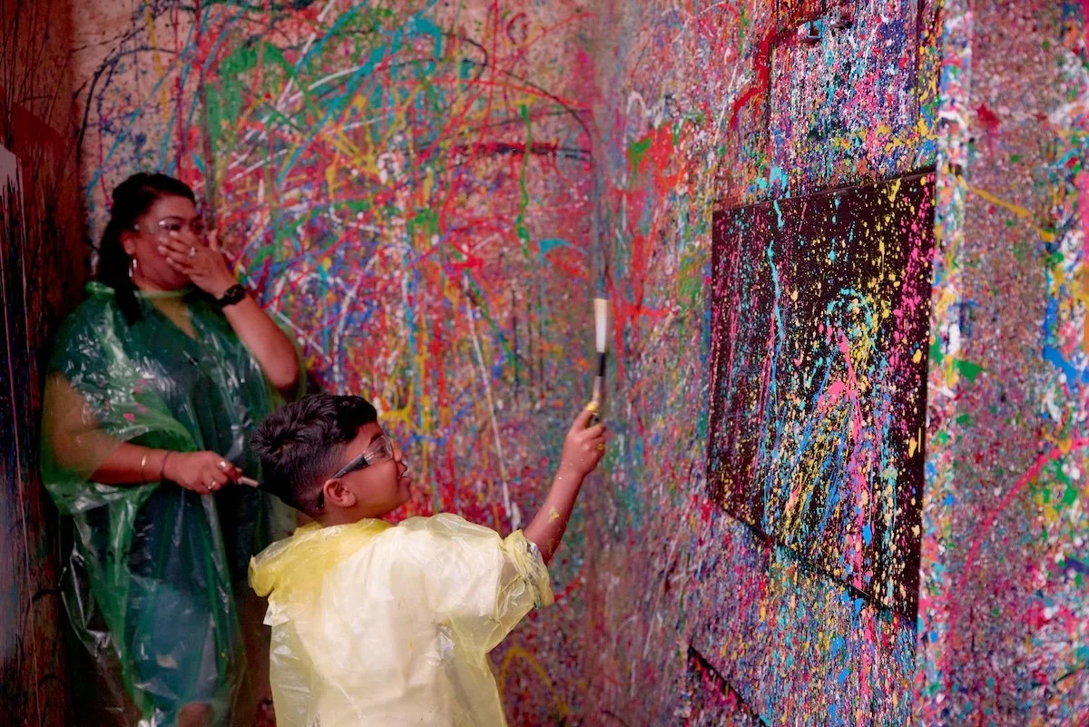 A woman and a young boy in a splattered paint splatter art room, with the boy using a paint scraper on the wall while the woman watches. The wall is covered in colorful splatters and abstract art.