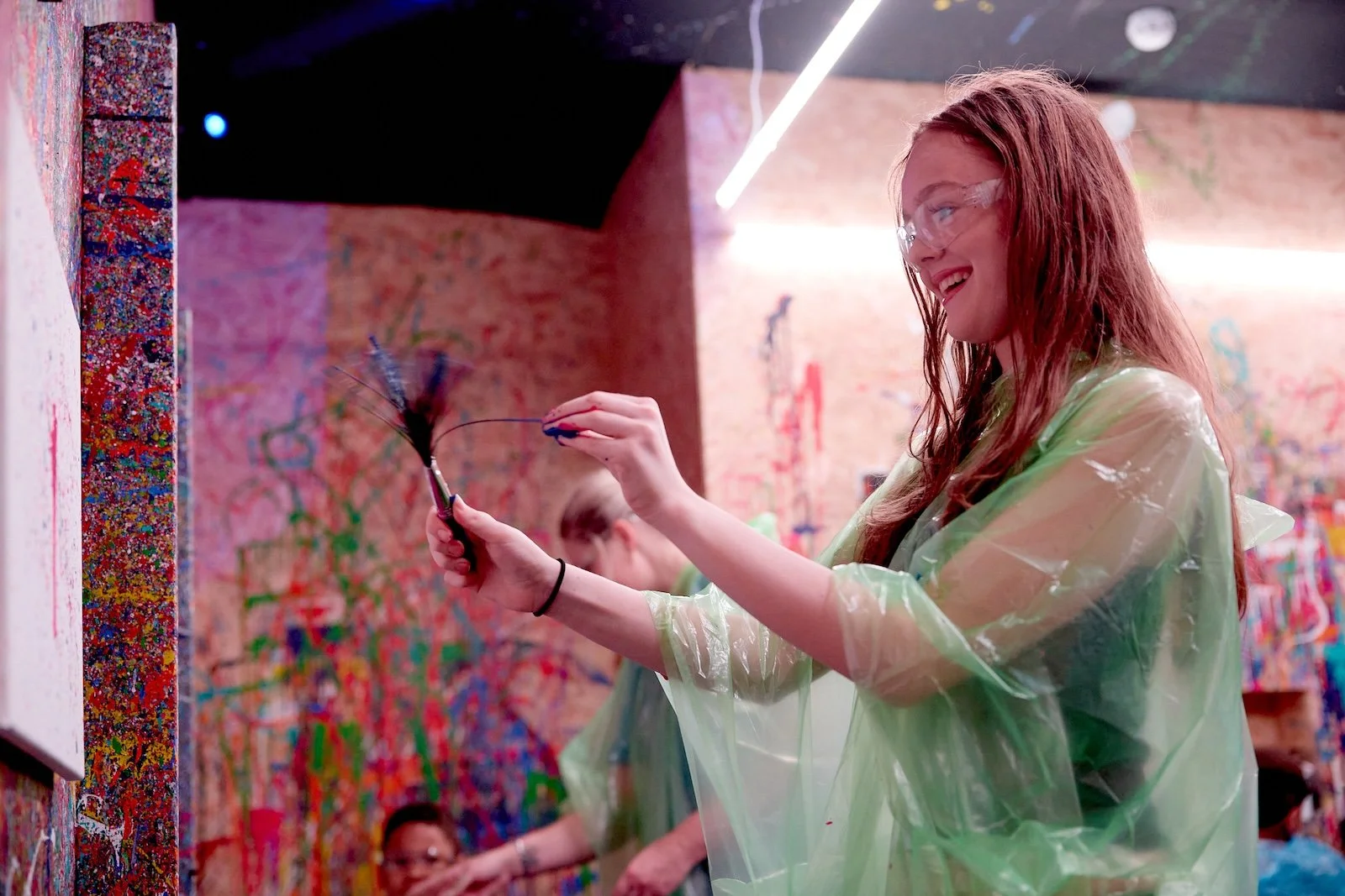 A woman with red hair and glasses smiling as she uses a feather duster in an art studio with colorful, abstract wall art.